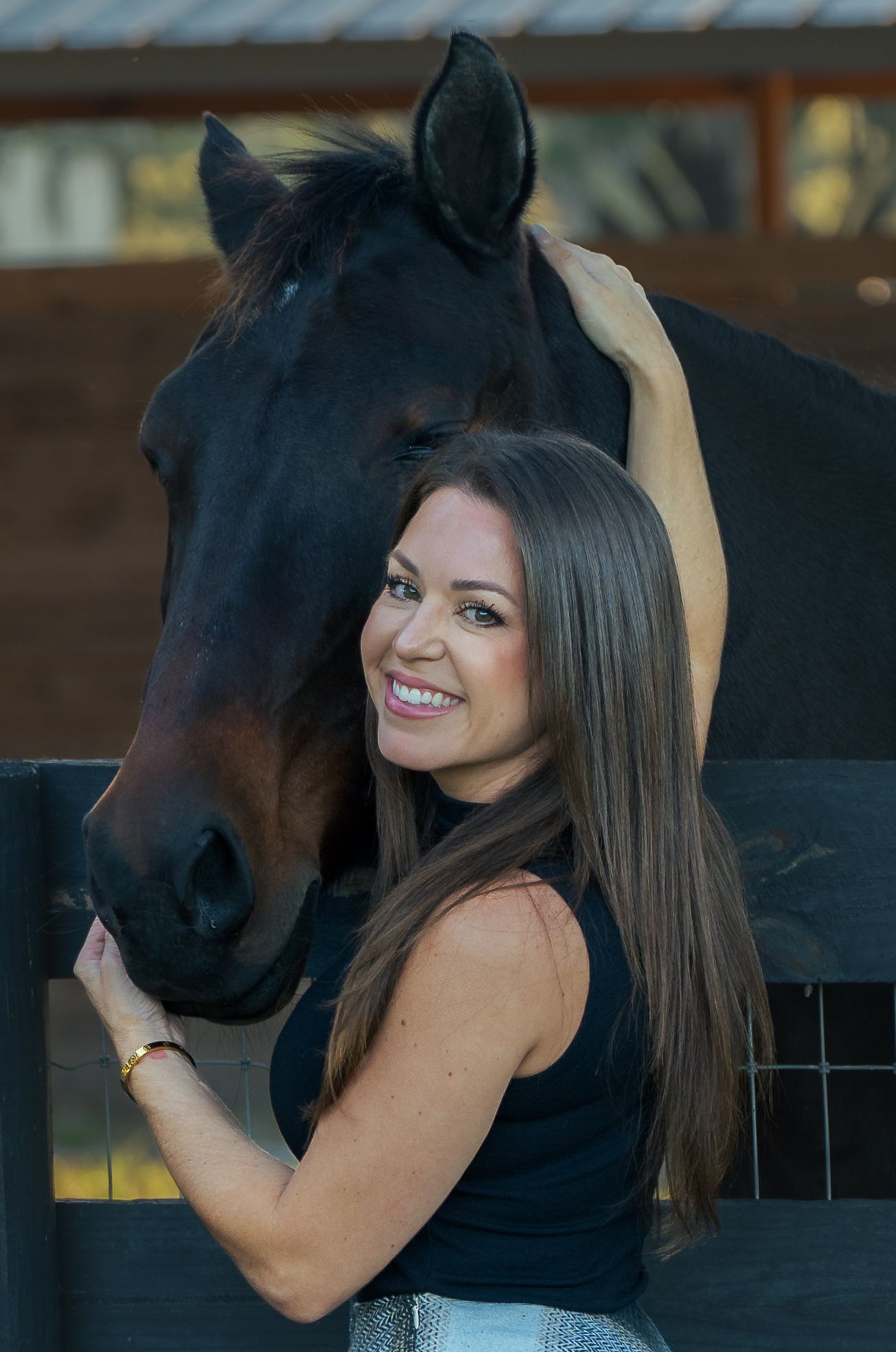 A smiling young woman with long brown hair embraces a black horse at an outdoor stable fence.