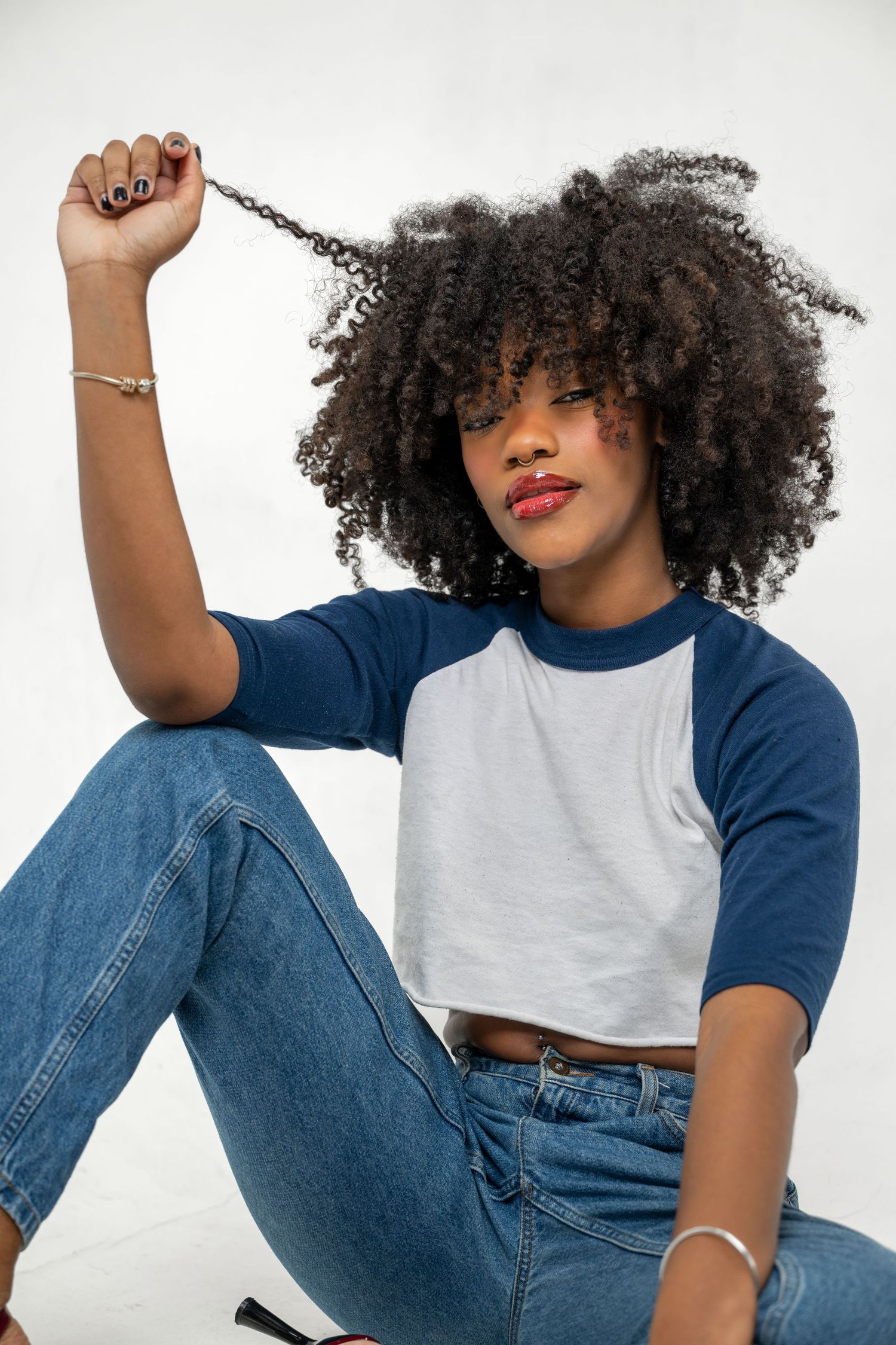 Black woman with natural afro hair holding a curl up while seated, wearing blue and white raglan crop top.