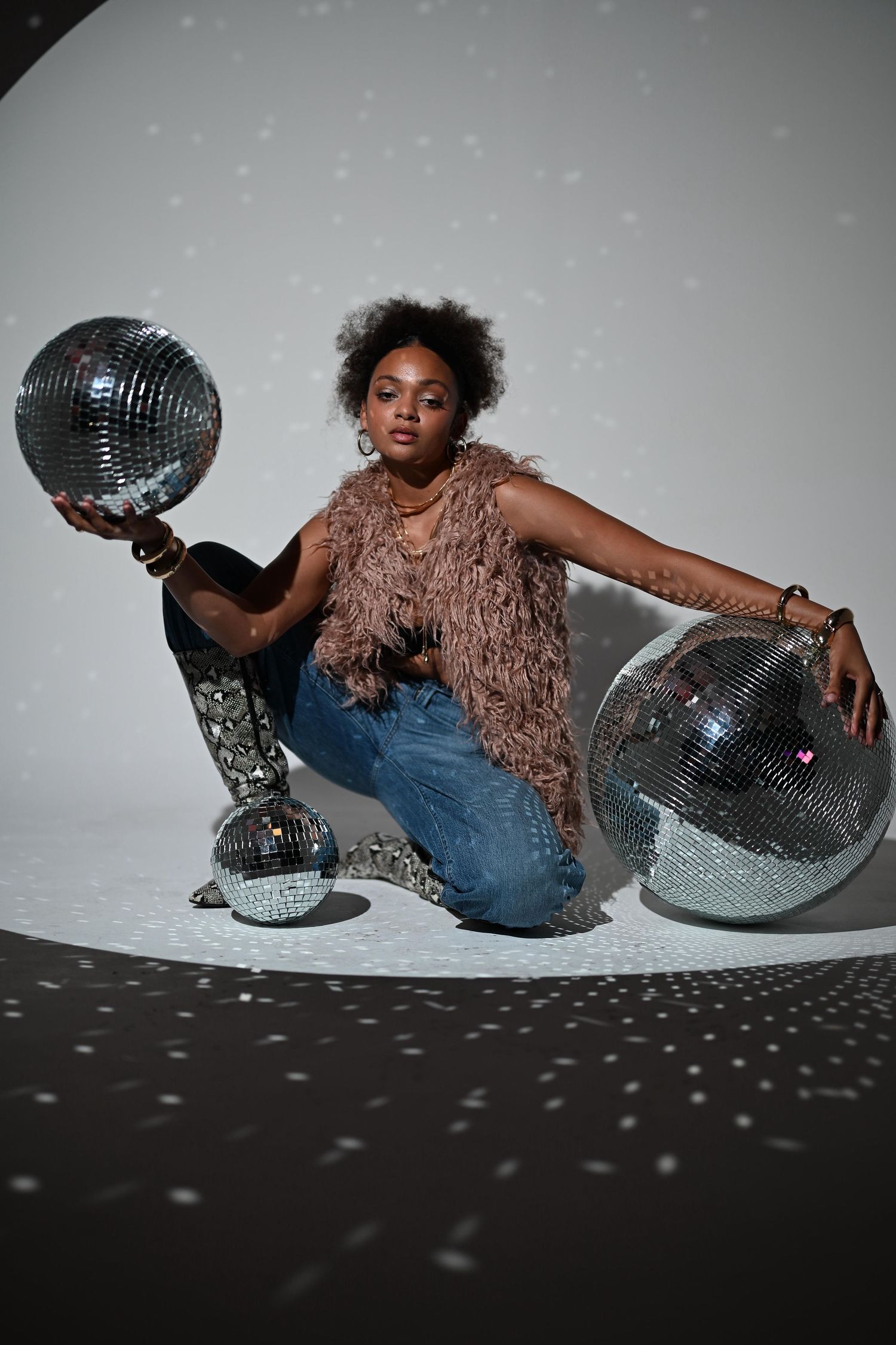 Black woman in fur vest and jeans kneels between glittering disco balls in dramatic studio lighting.