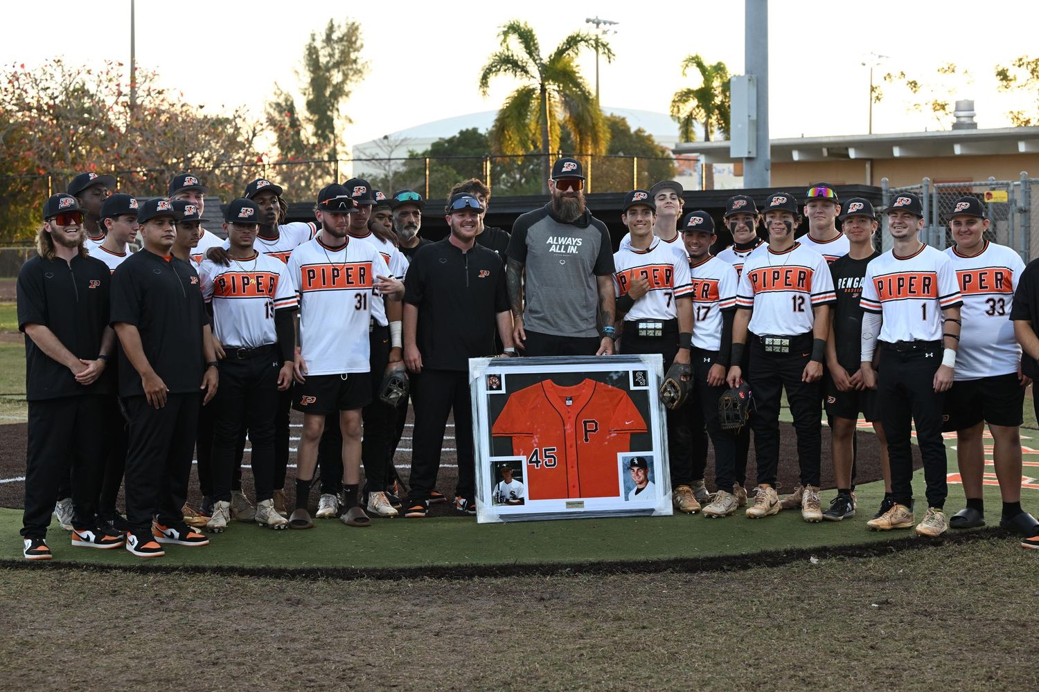 Baseball team poses with framed jersey during special recognition ceremony on outdoor field.