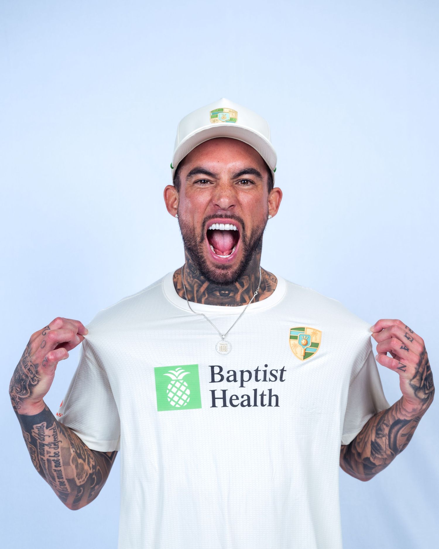 Tattooed man in white Baptist Health t-shirt and cap cheering with fists raised against light blue background.