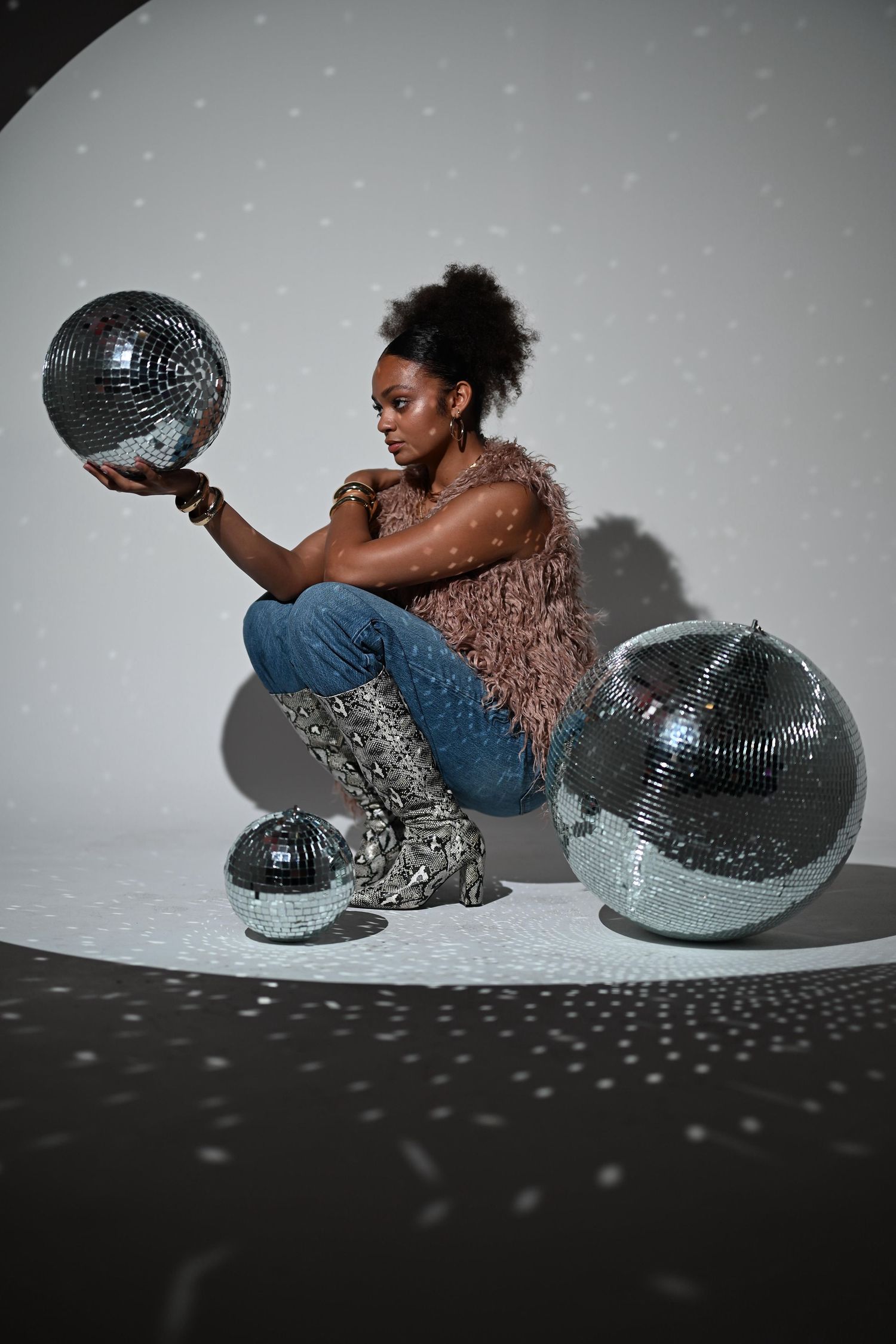 Black woman in pink fur vest and jeans poses with large silver disco balls in a spotlight studio setting.