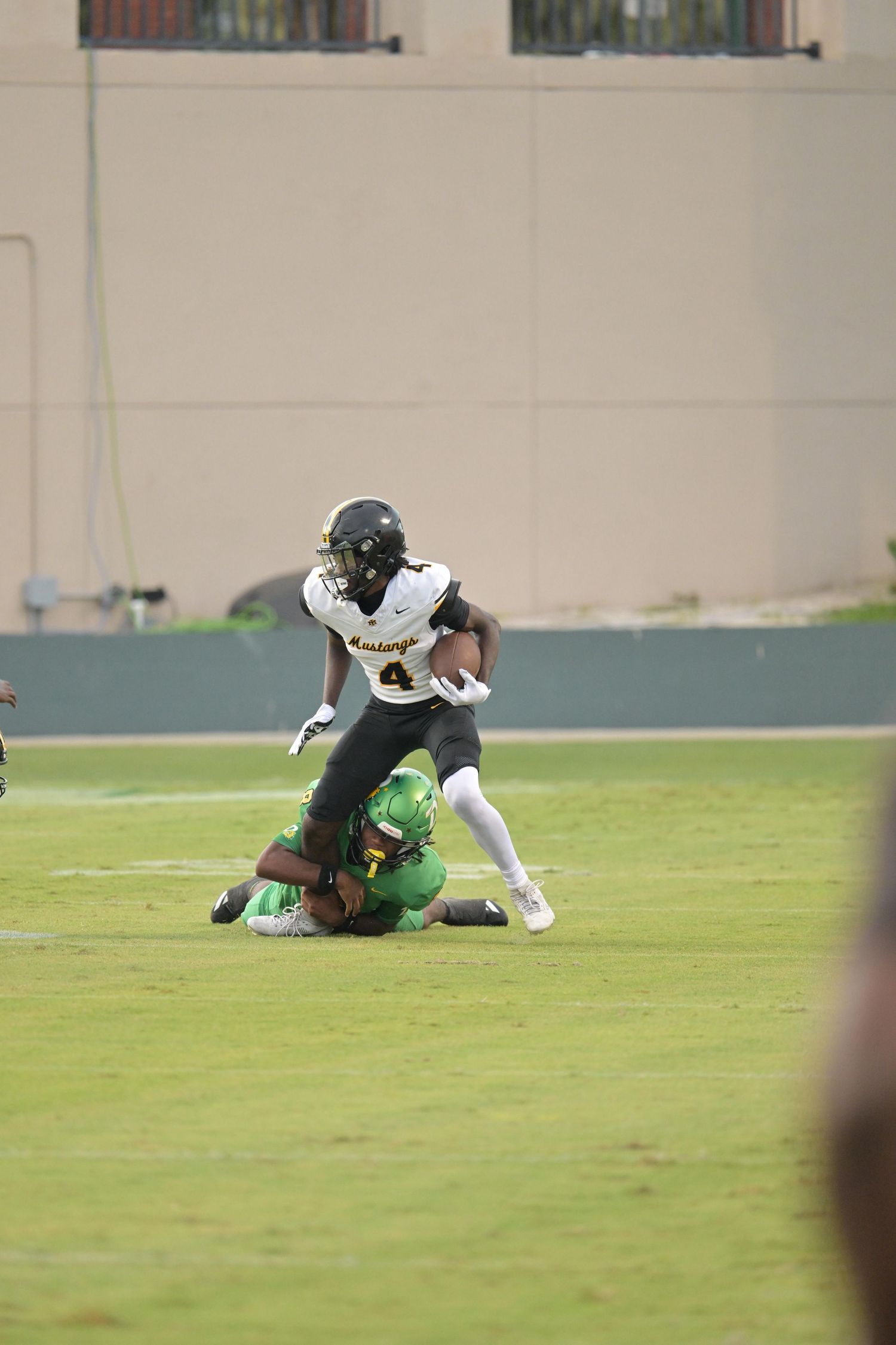 Football player #4 in black uniform carries the ball while being tackled by green-uniformed defender on grass field.