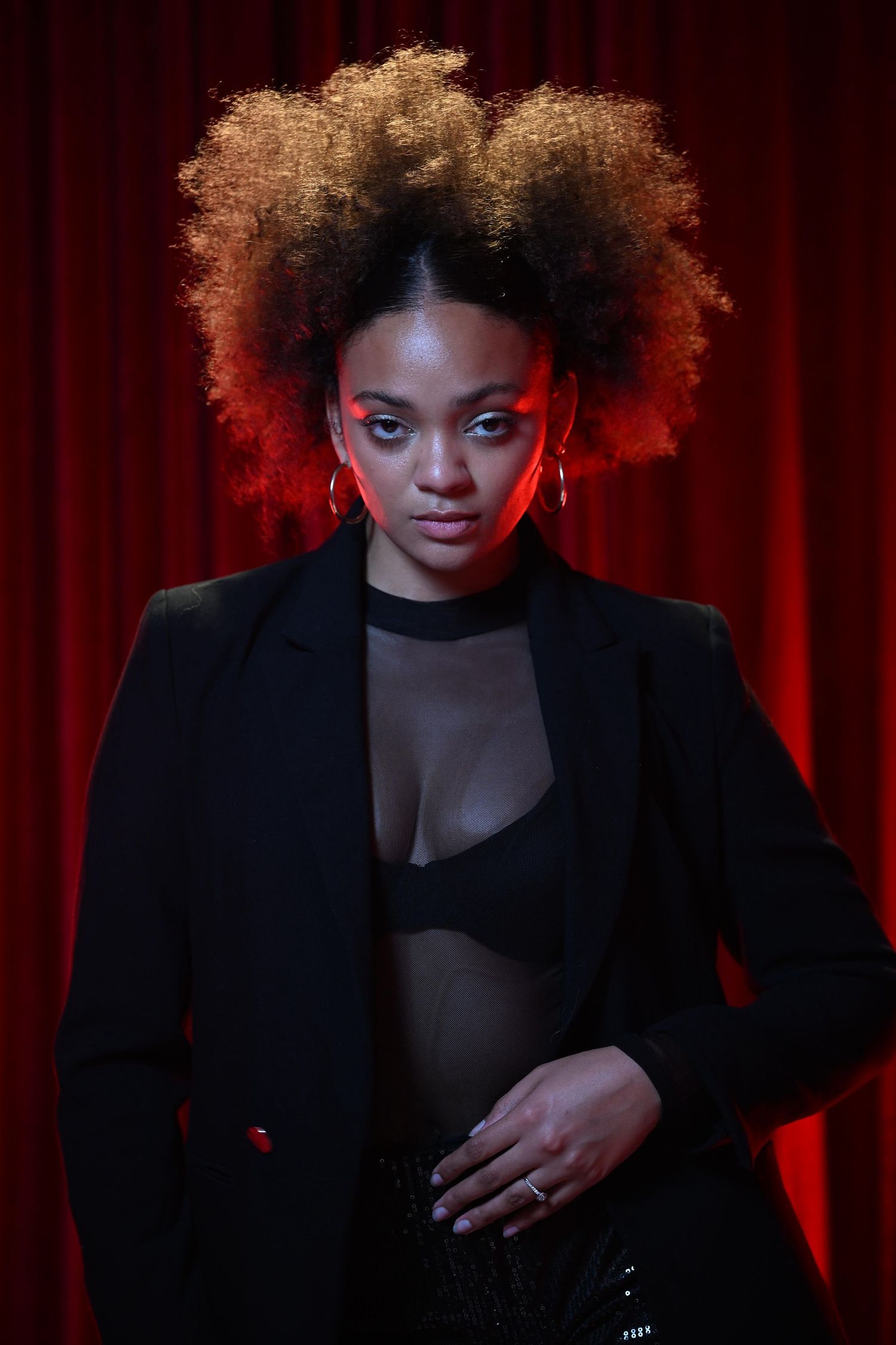 Stylish Black woman with afro hair in black blazer poses against red curtain in dramatic lighting.