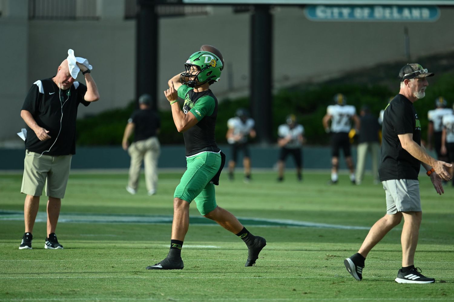 Football player in green uniform throws a pass during practice at City of DeLand stadium with coaches nearby.