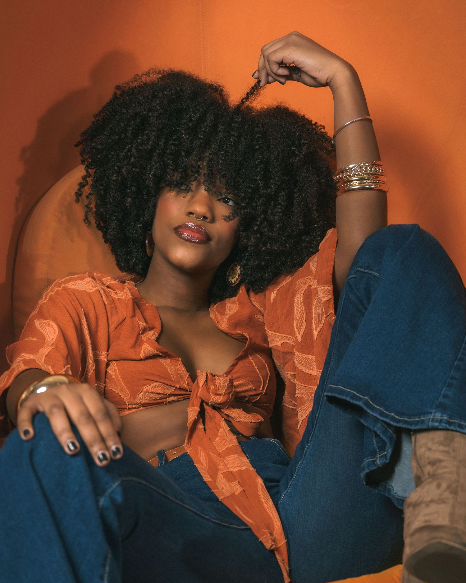 Young woman with natural afro hair wearing an orange tie-front crop top and blue jeans, posing in a chair.