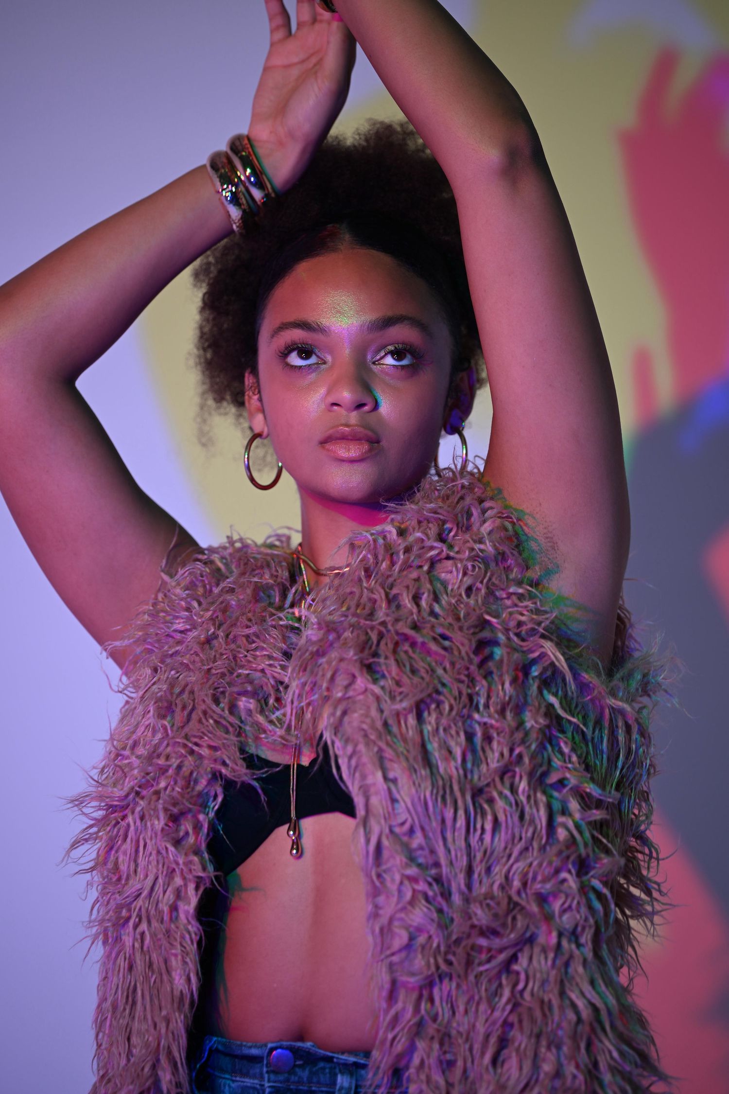 Black woman in pink fur vest raises arms under purple neon lights with glitter makeup and shadow art.