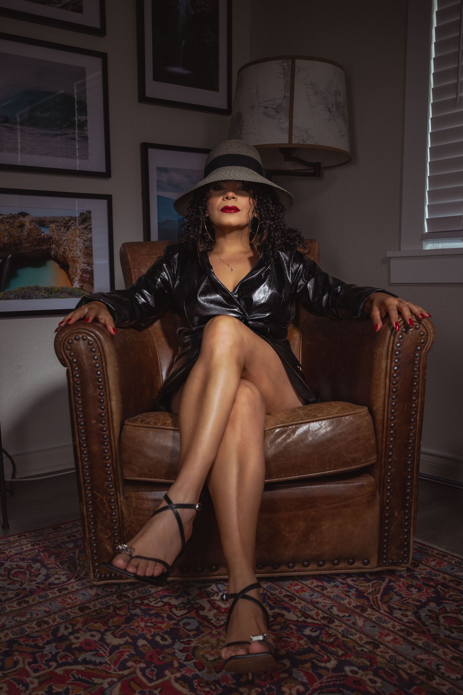 Woman in black jacket and fedora hat seated confidently in brown leather armchair on patterned rug.