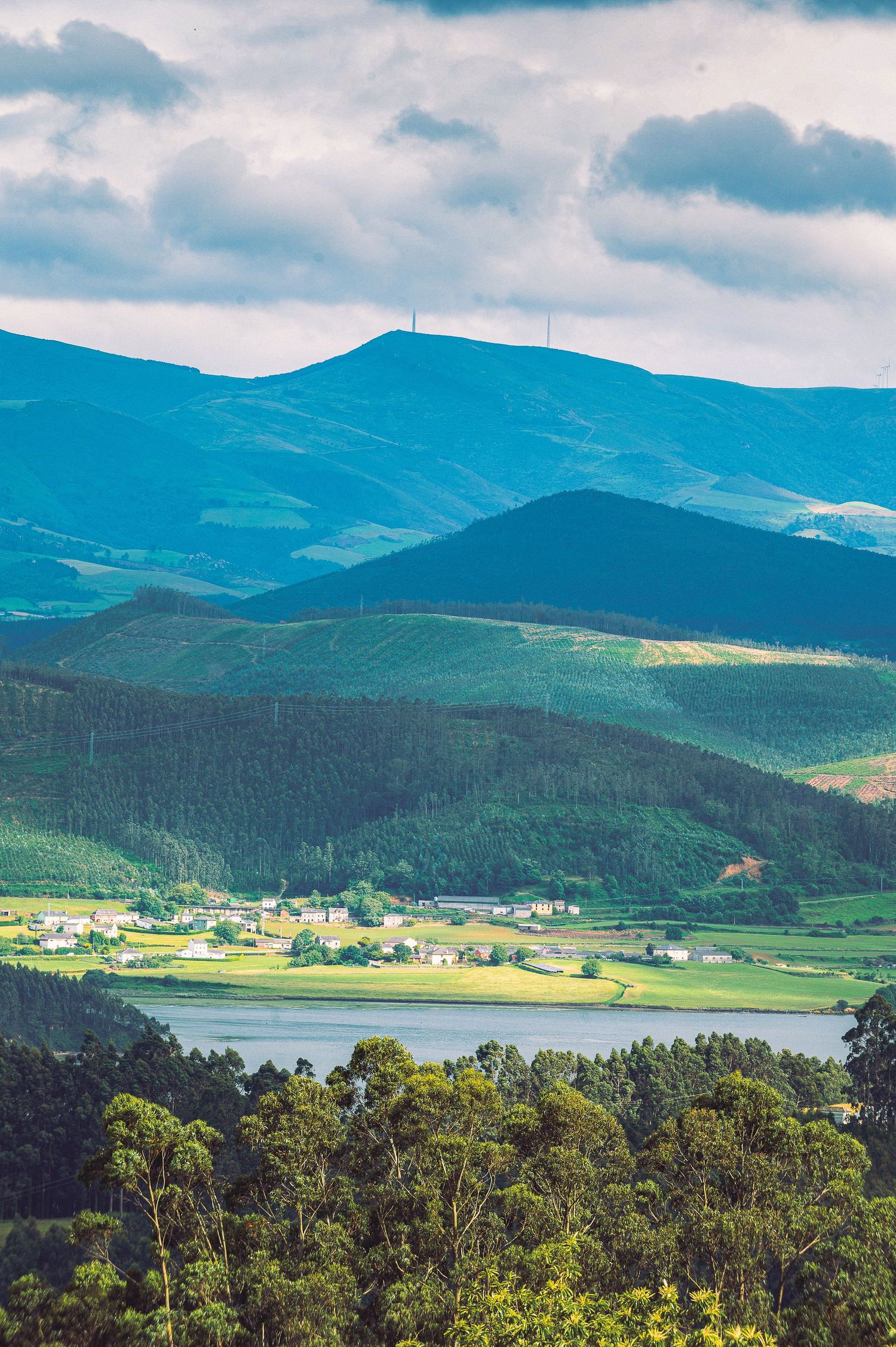 Aerial view of lush green valley with lake, small village, and mountain range under cloudy sky.
