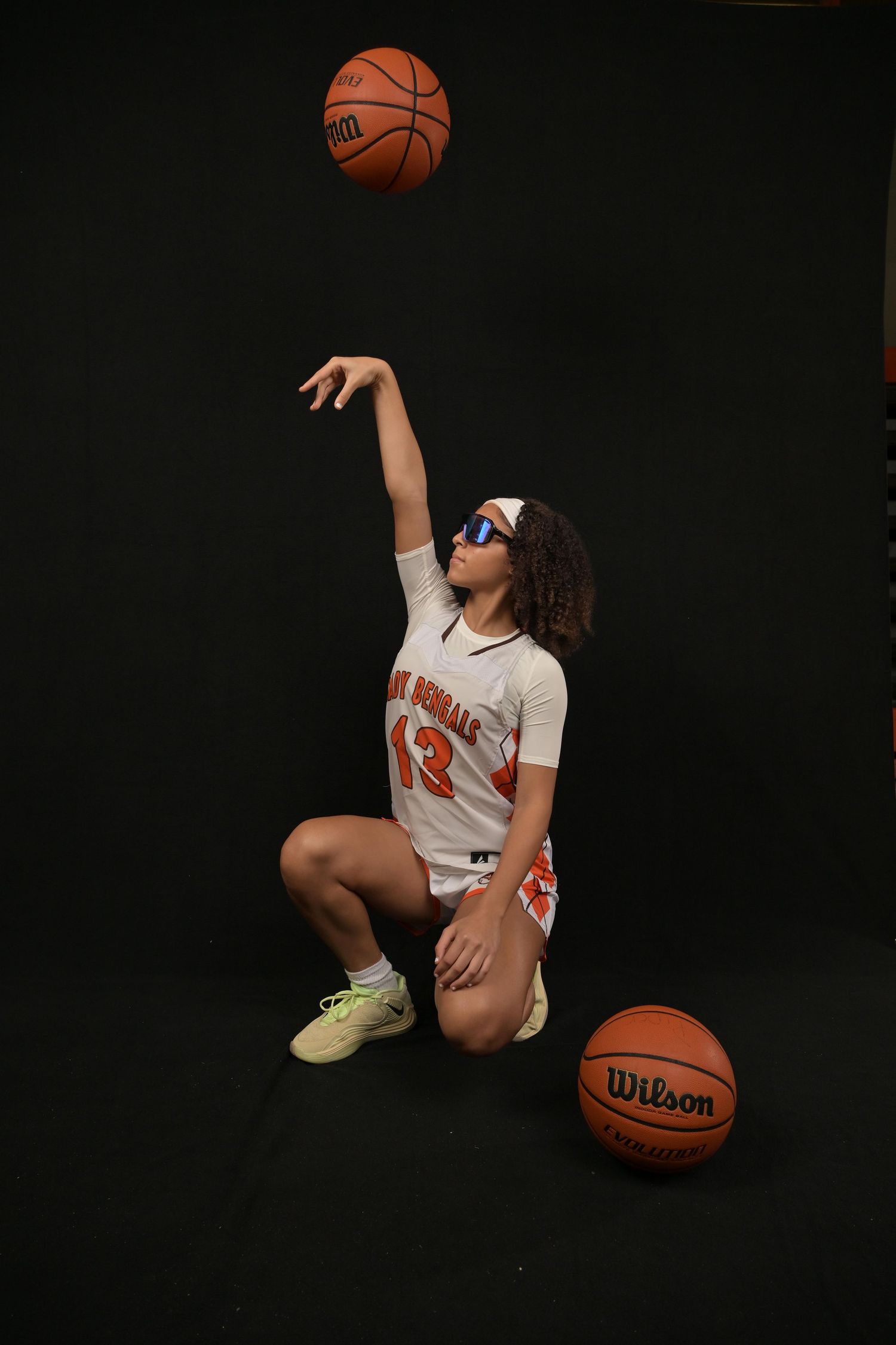 Basketball player in white jersey #13 kneeling, tossing a Wilson basketball up, with sunglasses on black background.