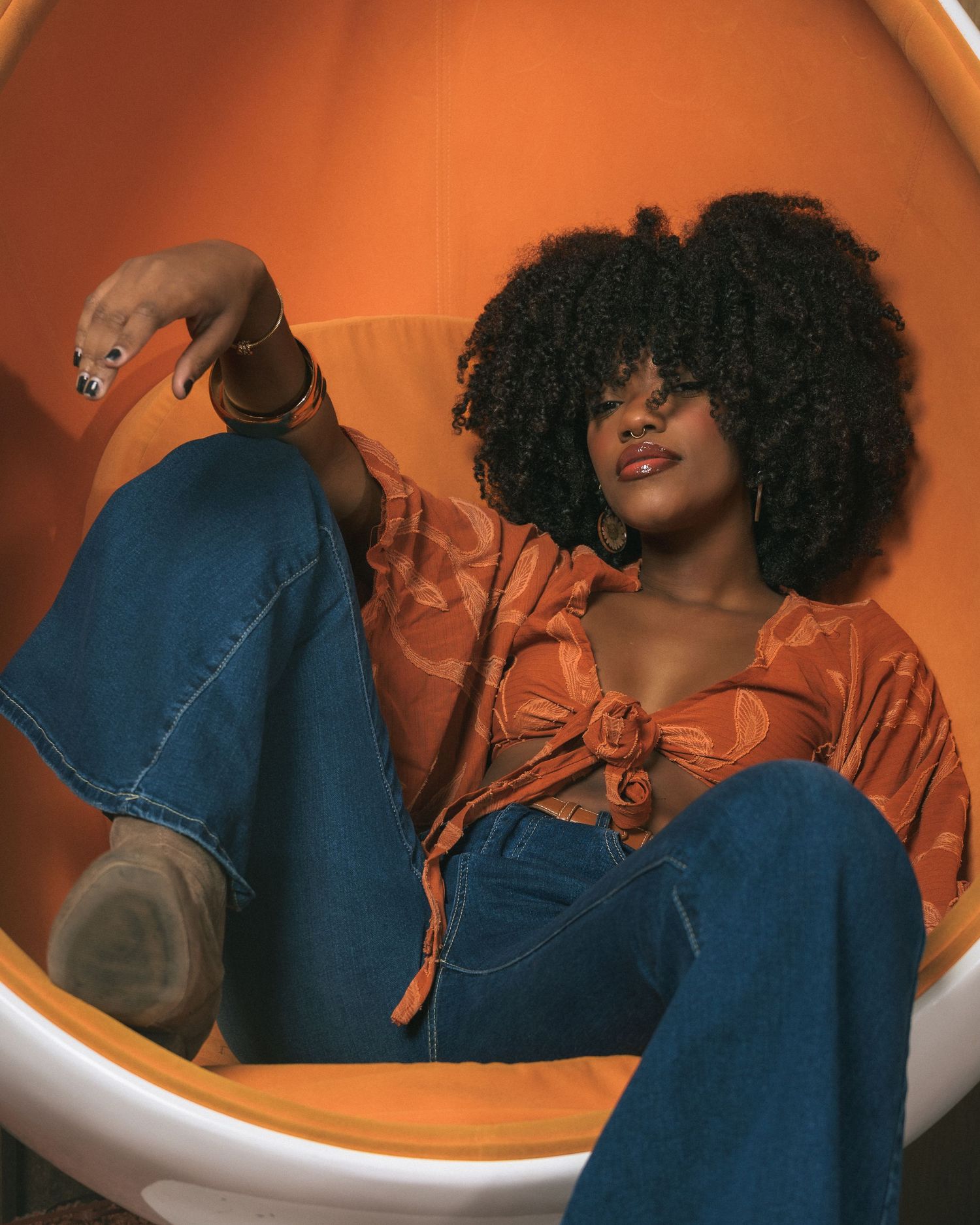 A fashionable woman with a natural afro sits in an orange egg chair wearing a tied orange top and wide-leg jeans.