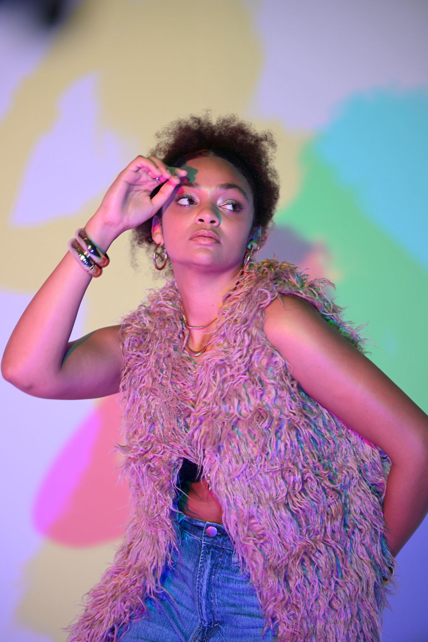 Black woman in pink fur vest poses under vibrant neon lights with colorful shadow projection on wall.