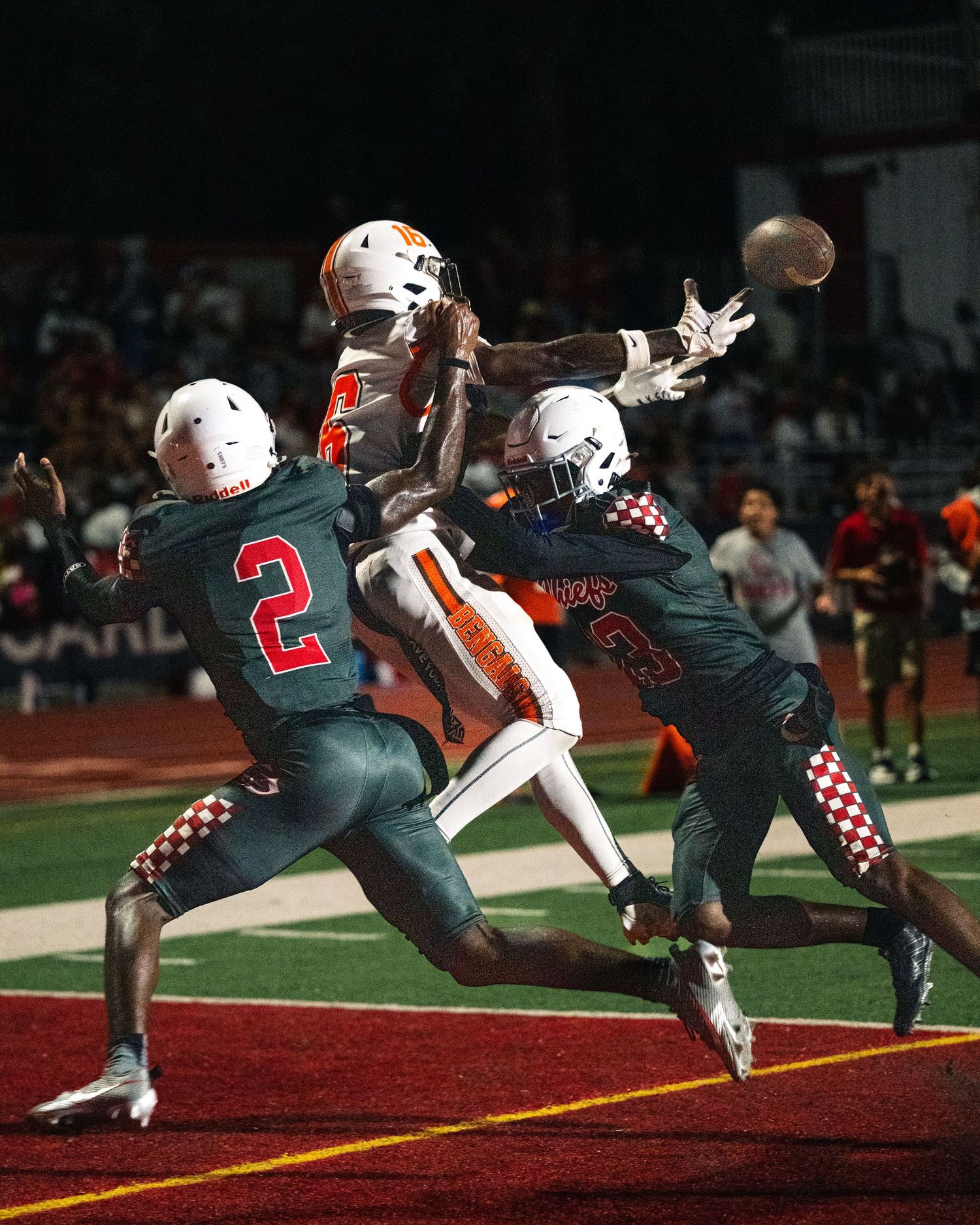 Football player in orange jersey leaping to catch a pass between two defenders in dark uniforms.