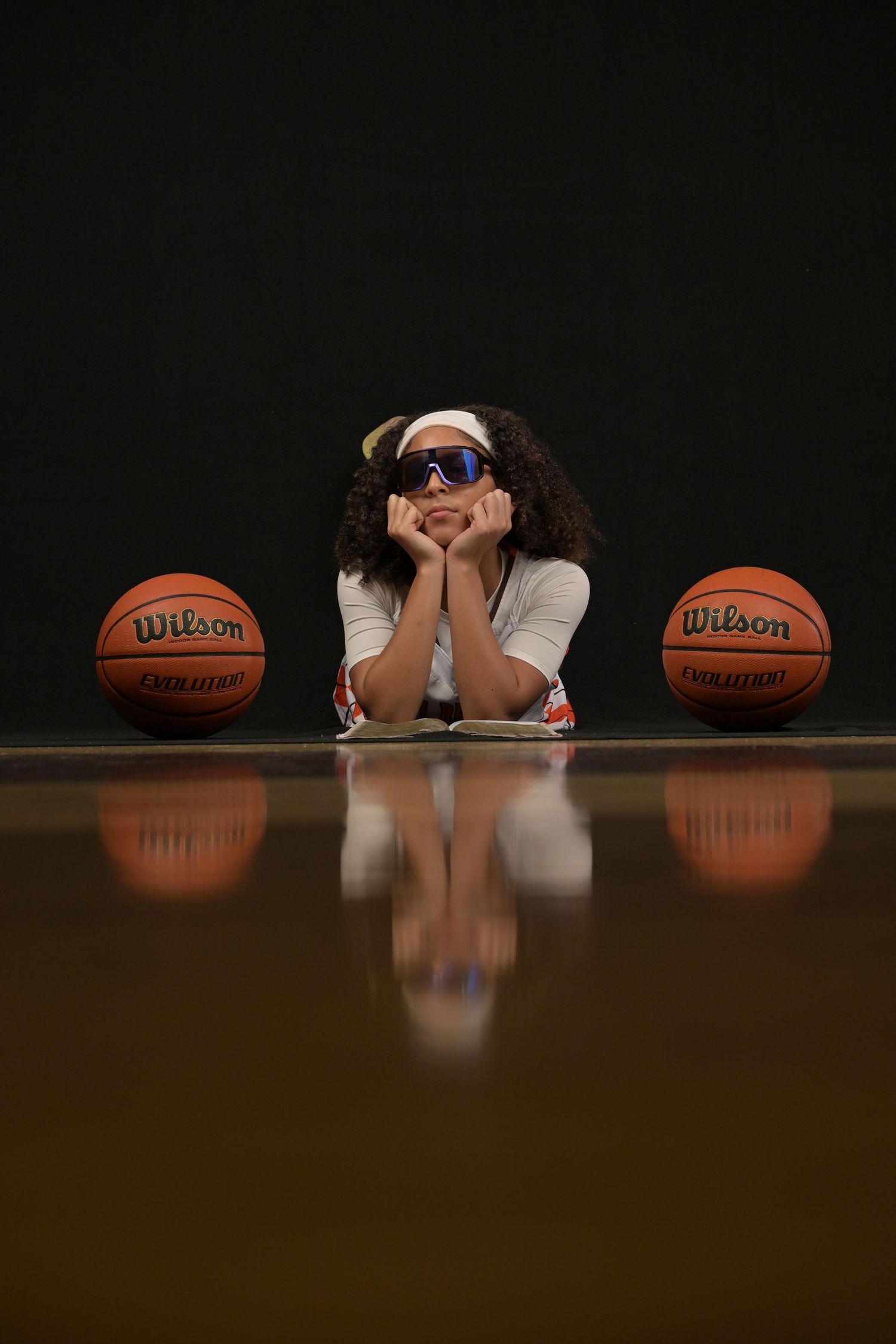 Young girl with curly hair and sport sunglasses lying on basketball court floor between two Wilson basketballs.