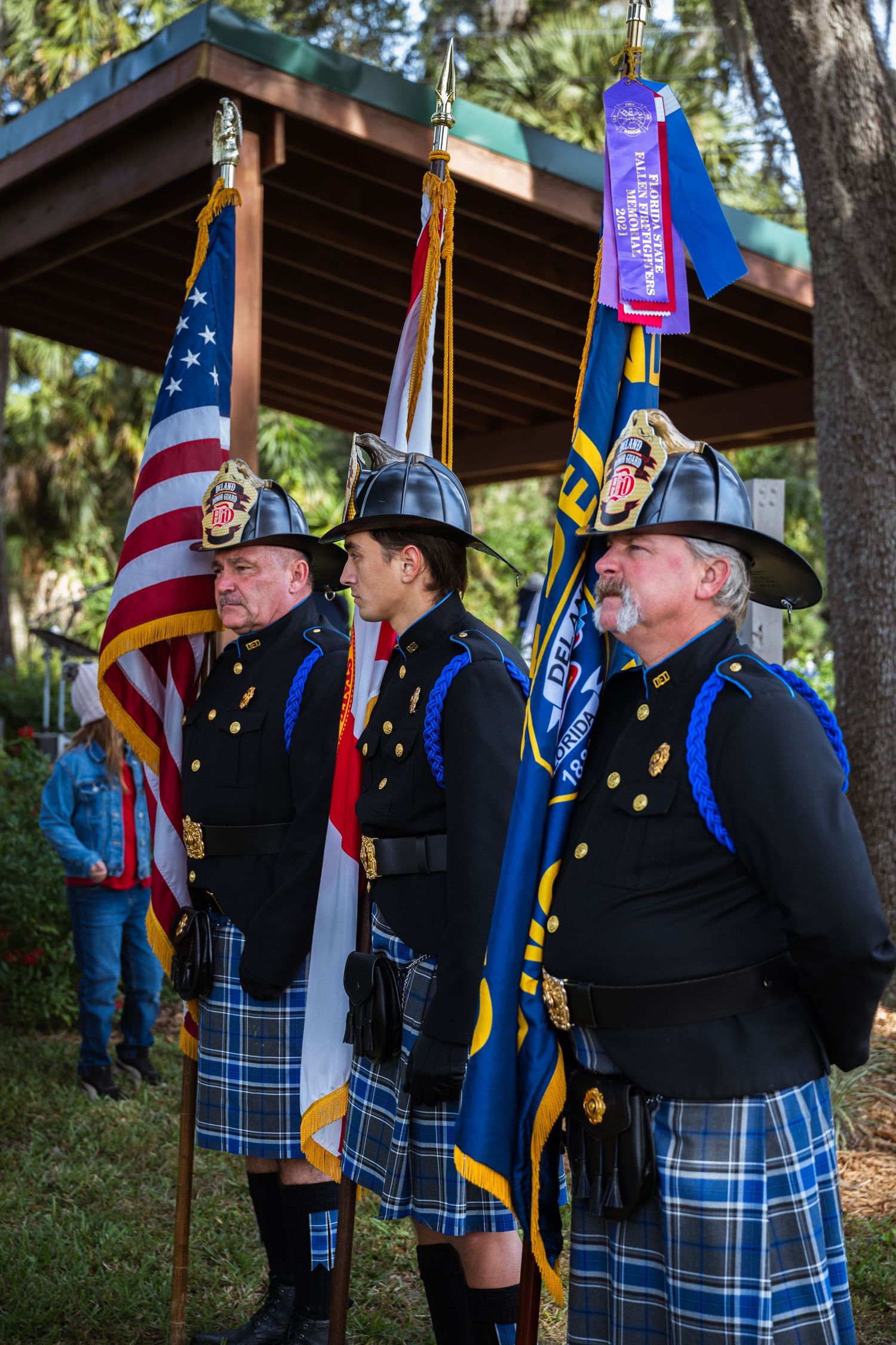 Three honor guard members in kilts and formal uniforms carry American and other flags at an outdoor ceremony.