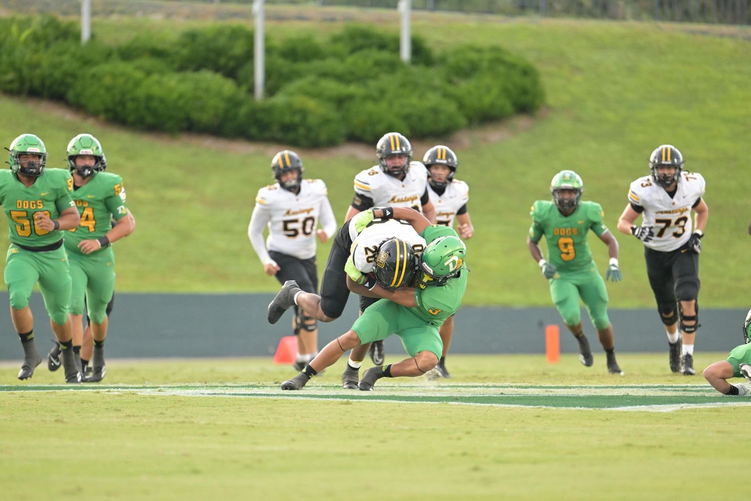 Green and white football players tackle an opponent during a high school football game on a well-maintained turf field.
