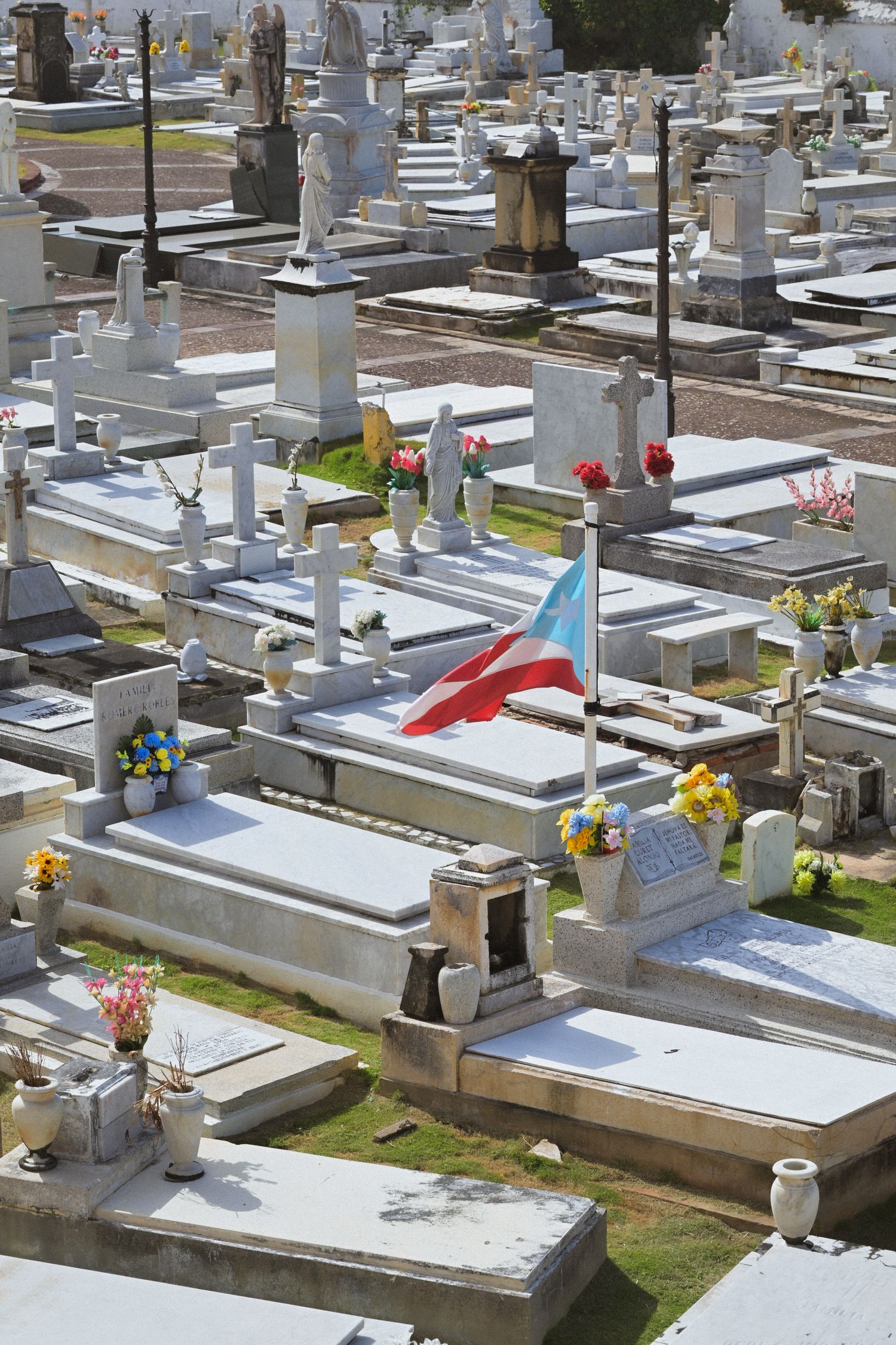 Puerto Rican flag among white tombstones and colorful flowers in sunlit cemetery grounds.