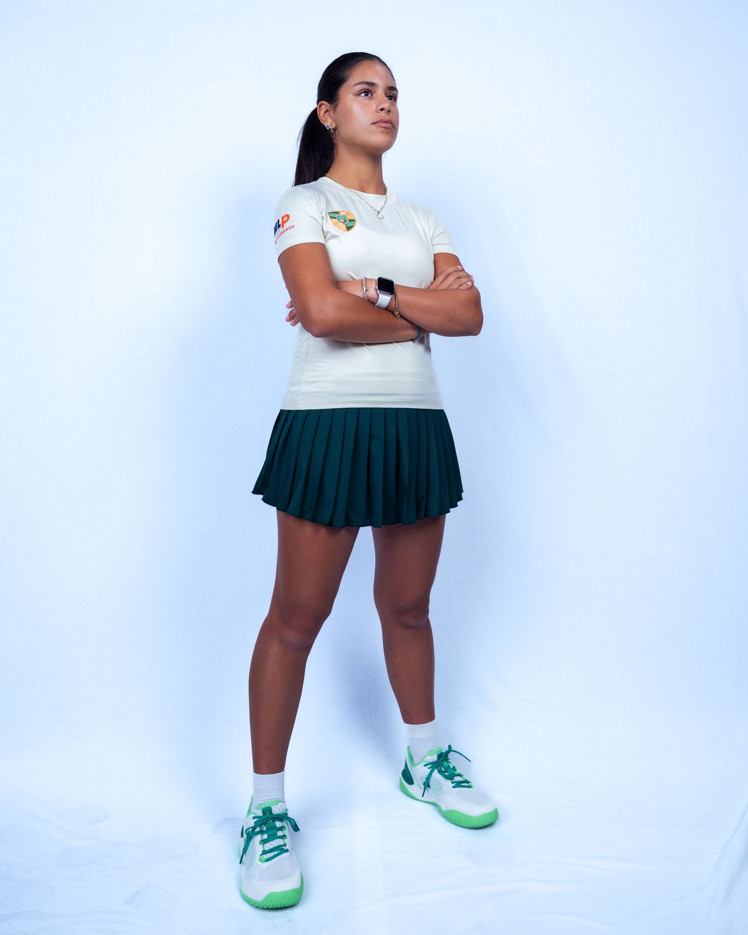 Female athlete in white top and green pleated skirt stands confidently with arms crossed wearing green and white sneakers.