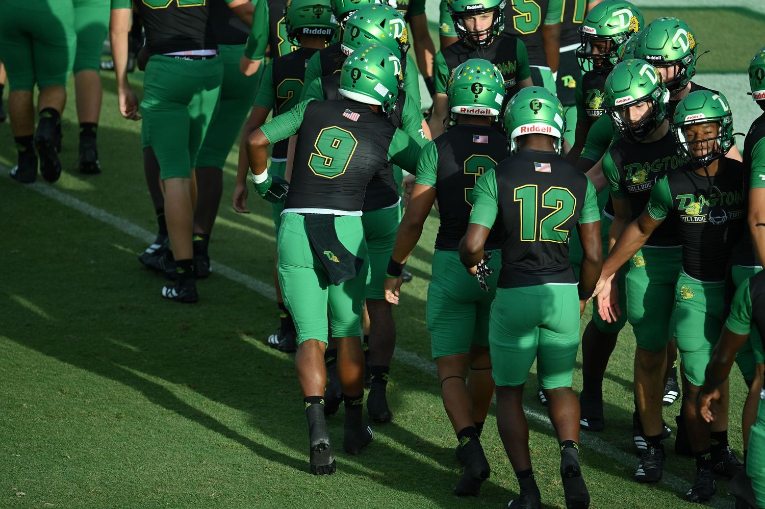 Green uniformed football players wearing numbers 9 and 12 huddle together on the field during a game.