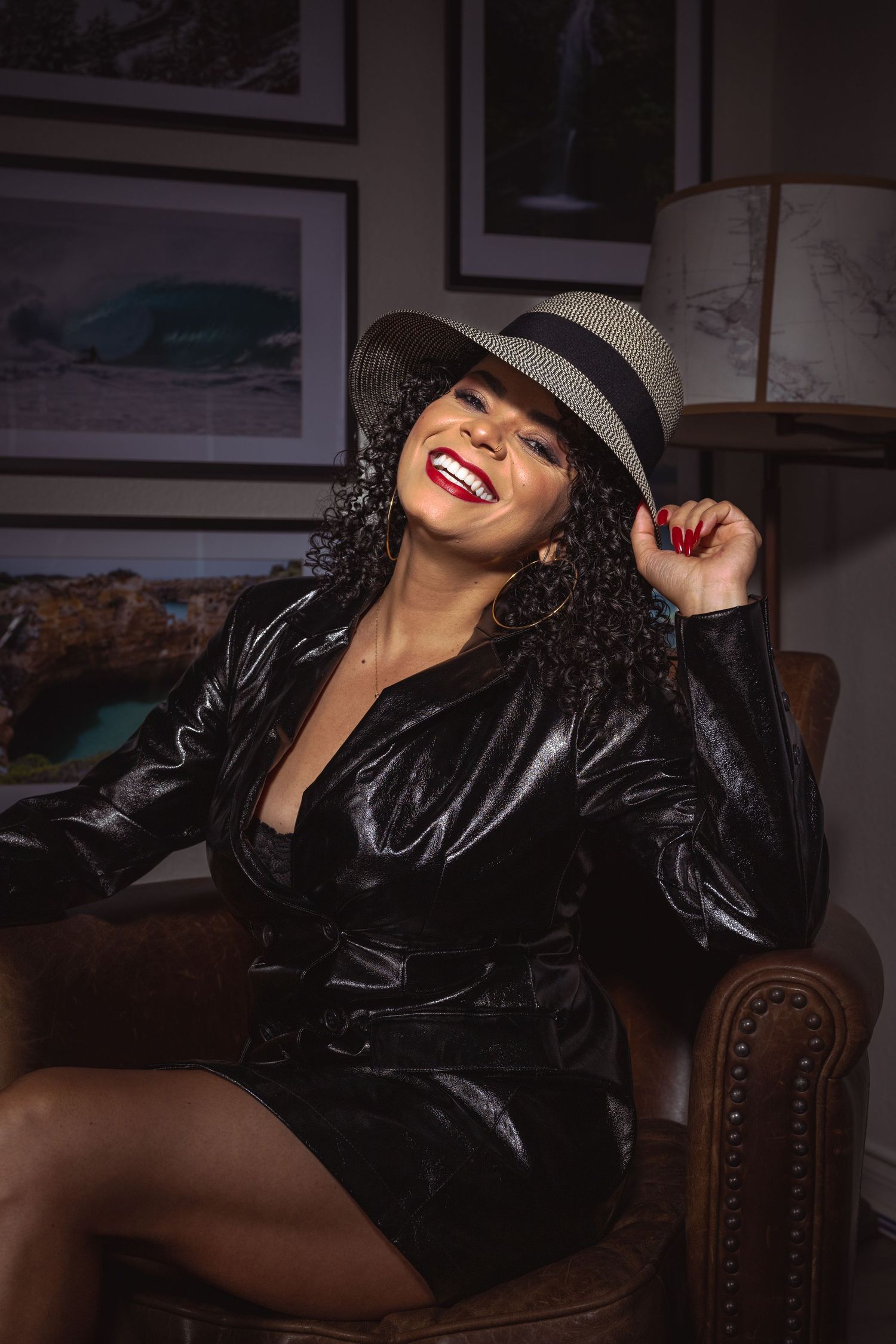 Smiling woman in black leather dress and fedora hat sitting in leather chair with framed art behind her.