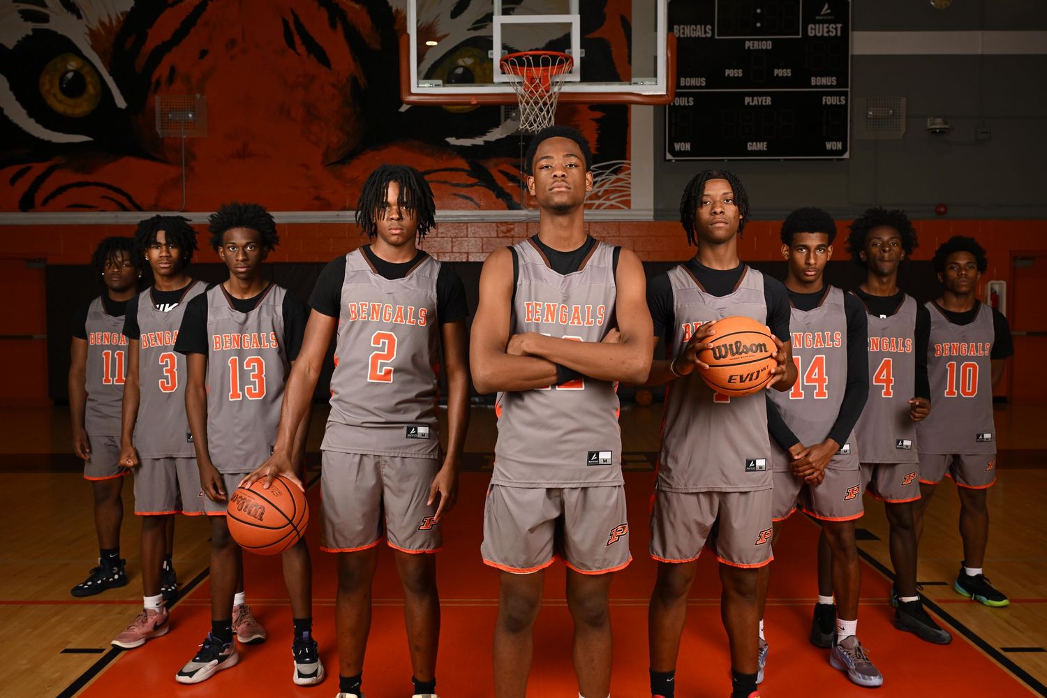 High school basketball team poses together in orange and gray uniforms inside gymnasium with tiger mural.