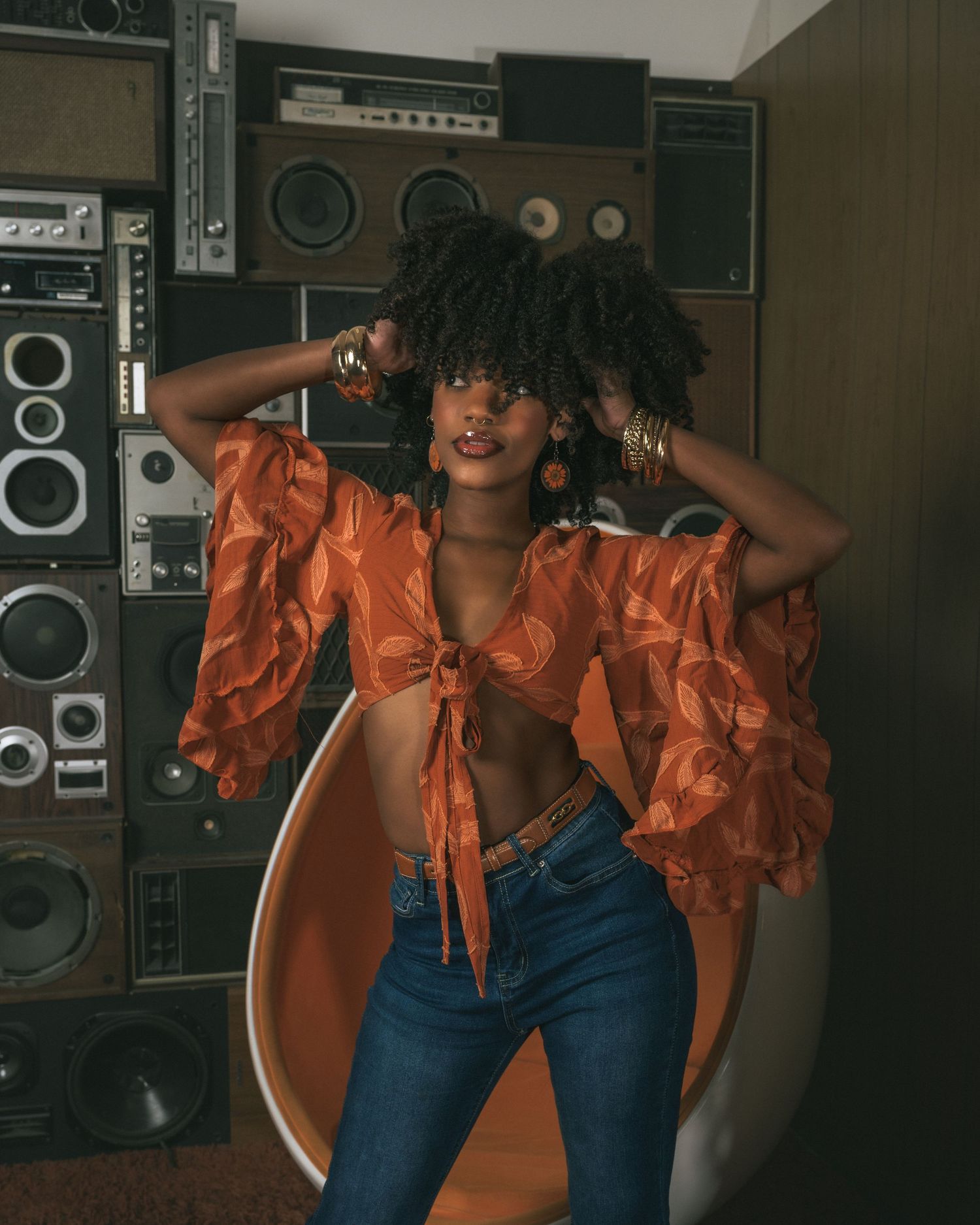 A stylish woman in an orange ruffled crop top and jeans poses in front of a wall of vintage stereo speakers.