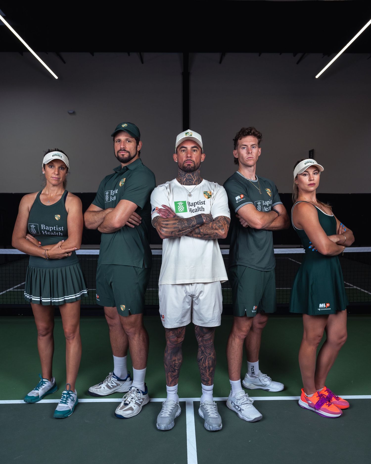 Five tennis players in green and white uniforms pose with arms crossed on an indoor tennis court.