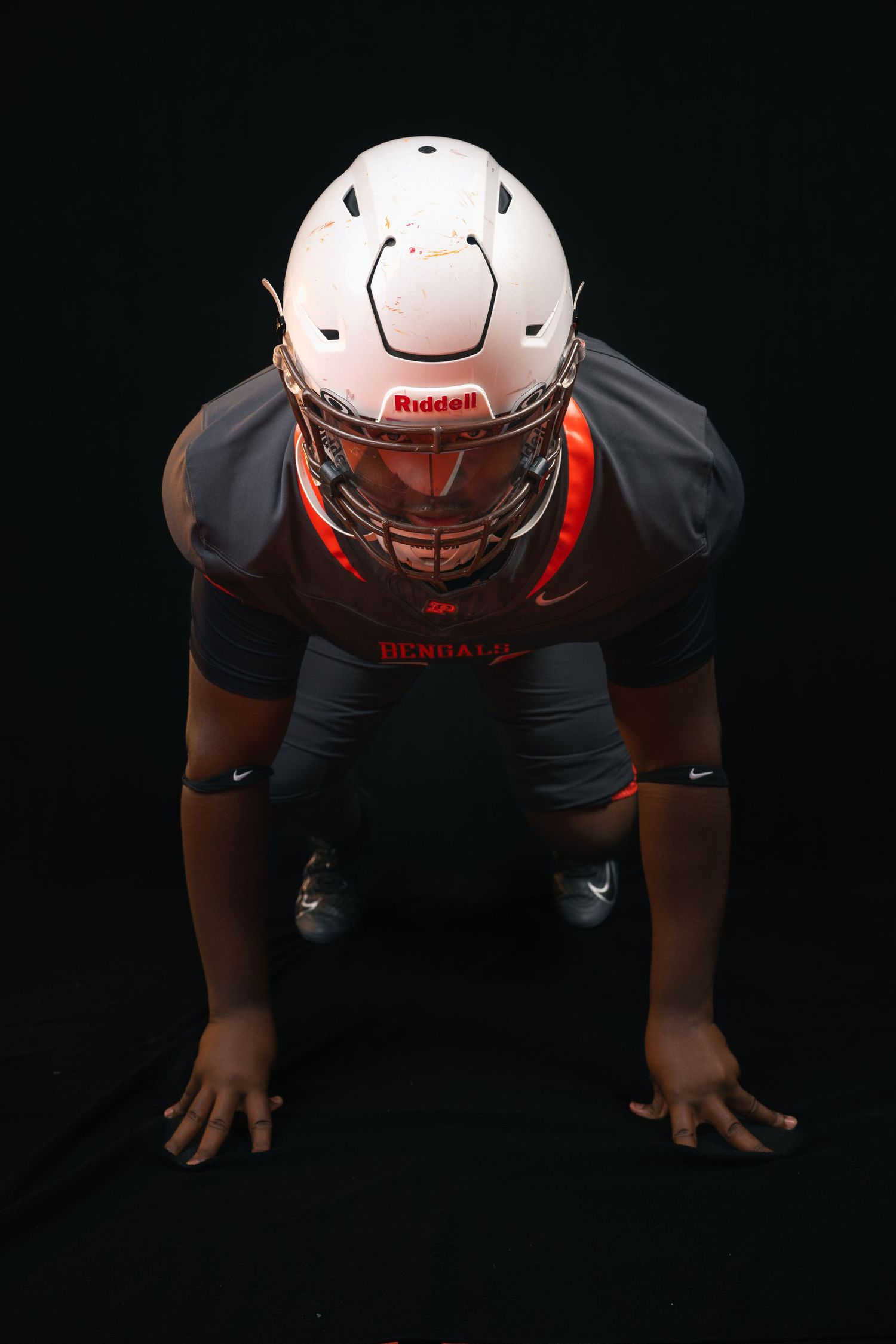 Bengals football player in white Riddell helmet crouched in three-point stance on black background.