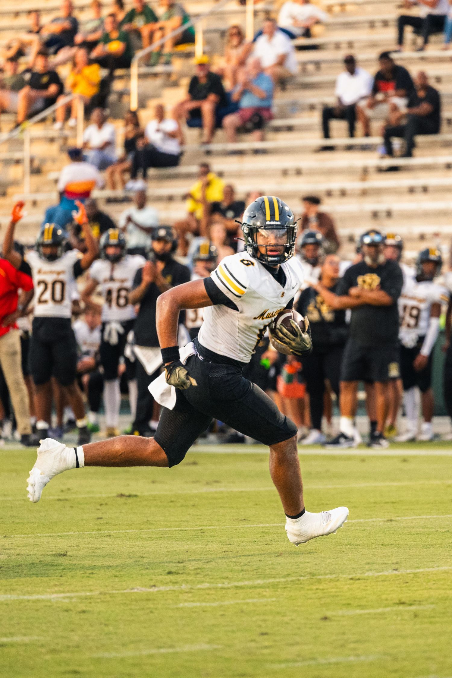A football player in black and gold uniform runs with the ball during a night game with fans in the stands.
