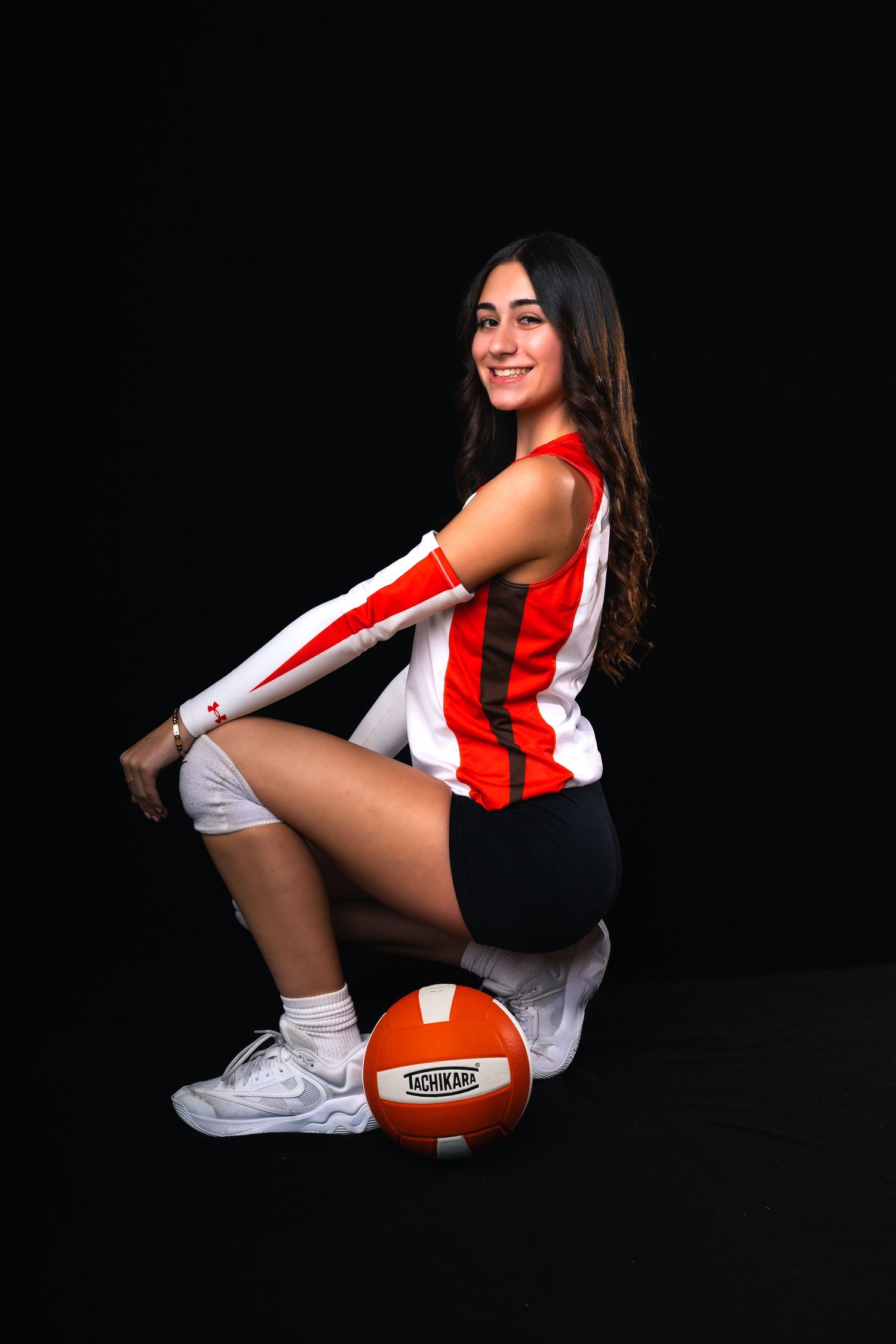 Young female volleyball player in red and white uniform sits on a Tachikara ball, smiling over her shoulder against black background.