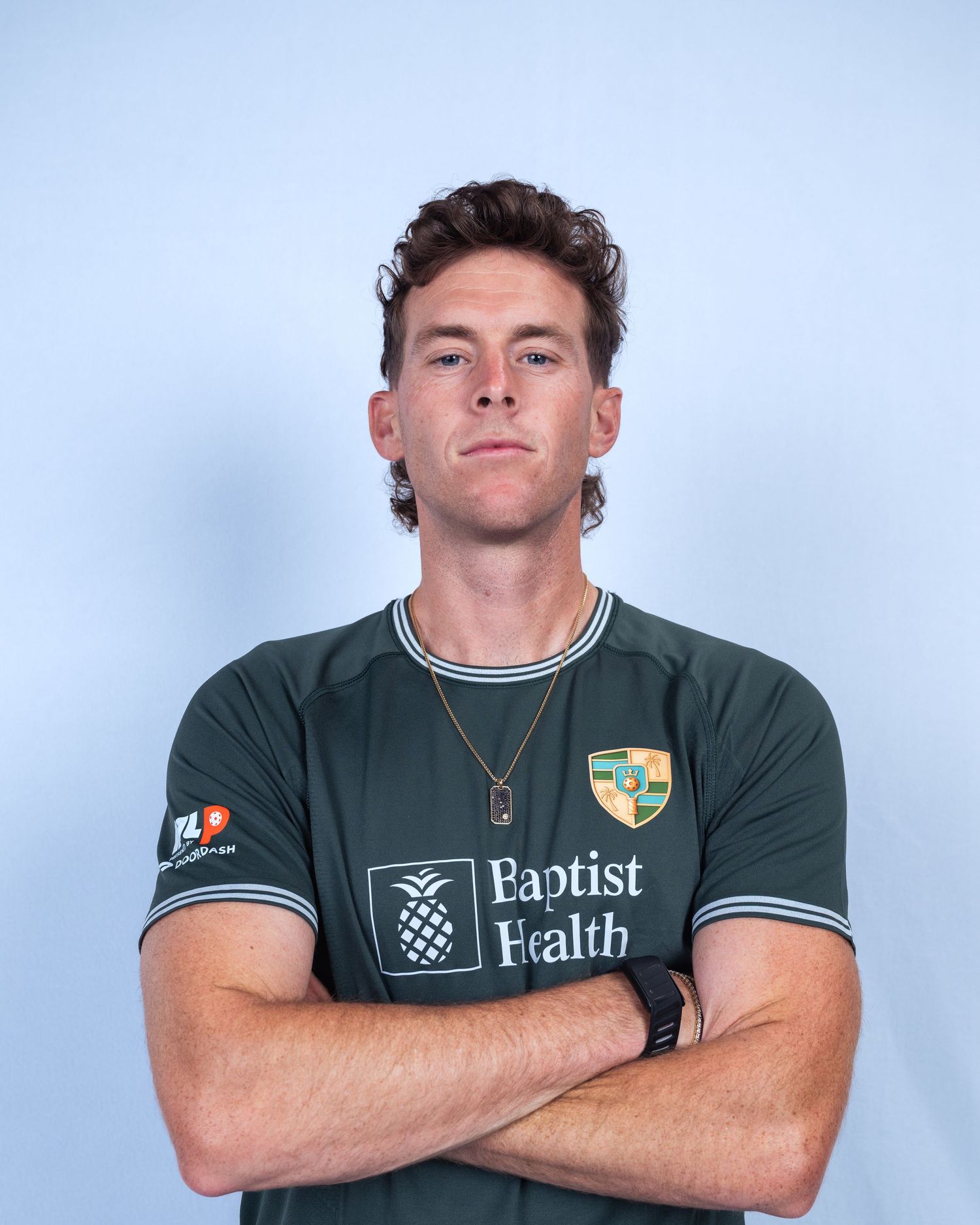 Young male athlete in dark green Baptist Health soccer jersey poses with arms crossed against light blue background.