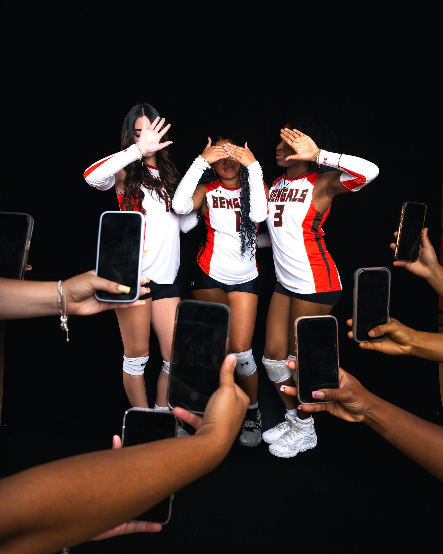 Three Bengals volleyball players cover their eyes while teammates photograph them with smartphones in a dark studio.