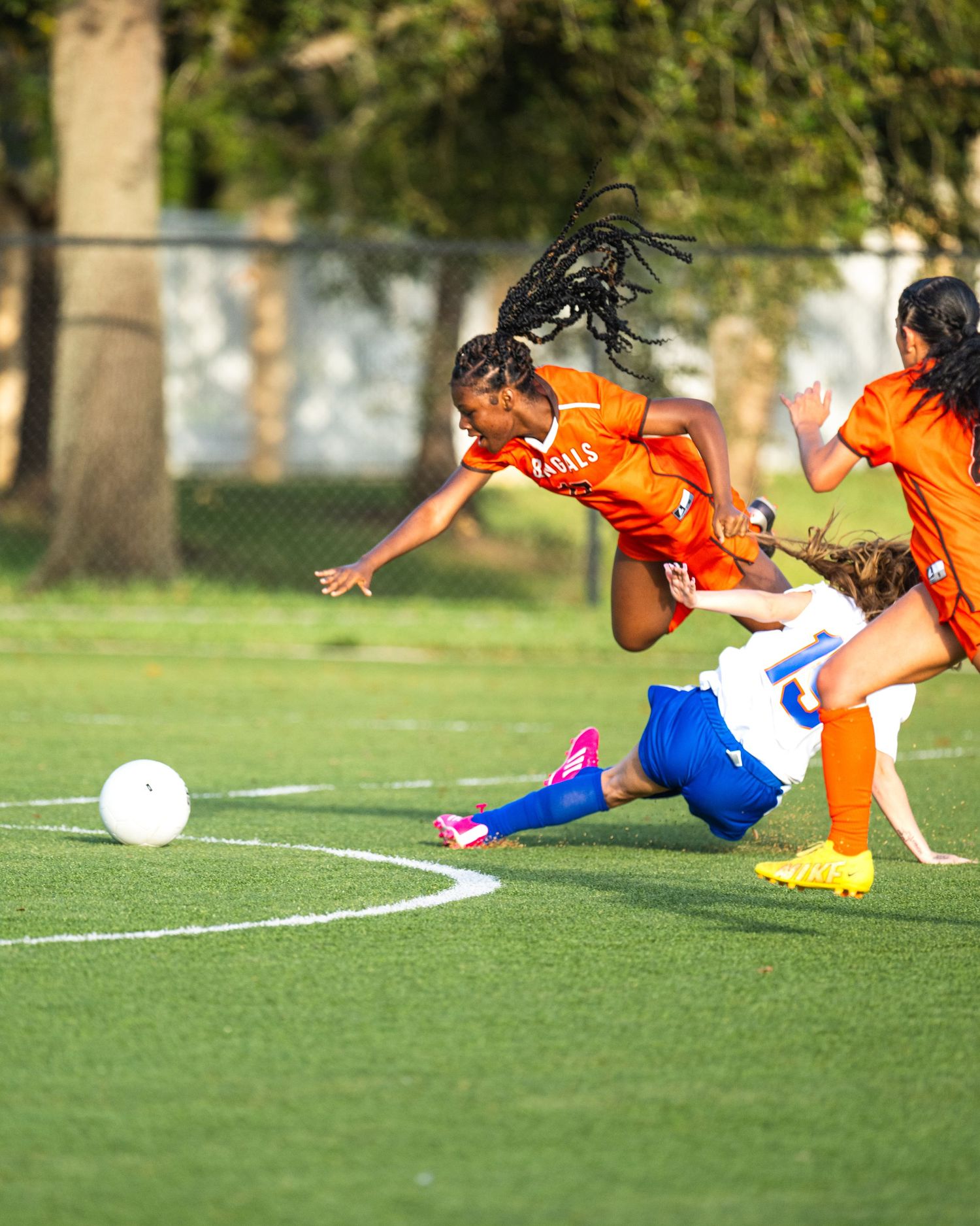 A female soccer player in an orange Bengals jersey leaps over a sliding opponent on a green field.