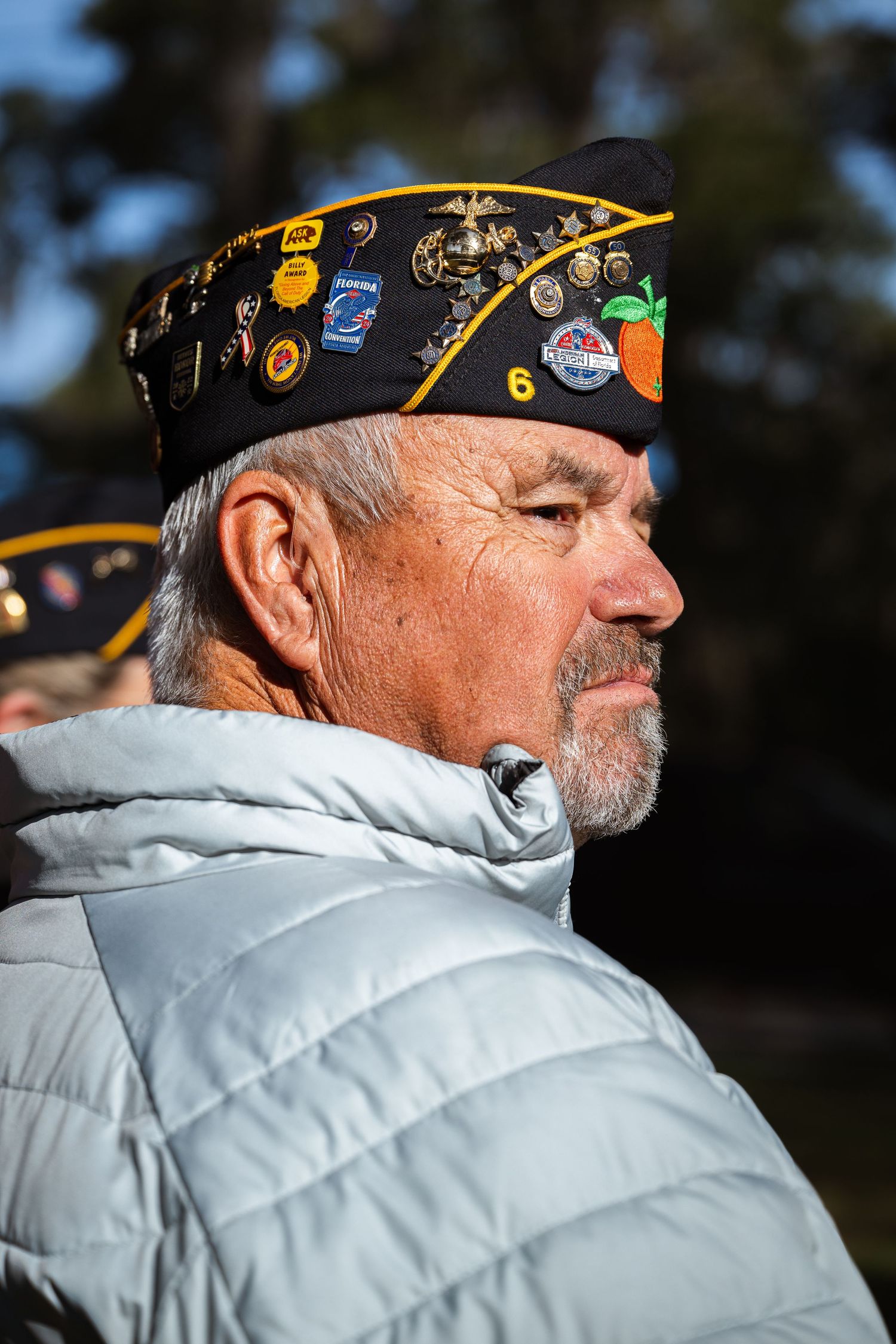 A decorated military veteran wearing a cap with pins and badges stands in profile outdoors.