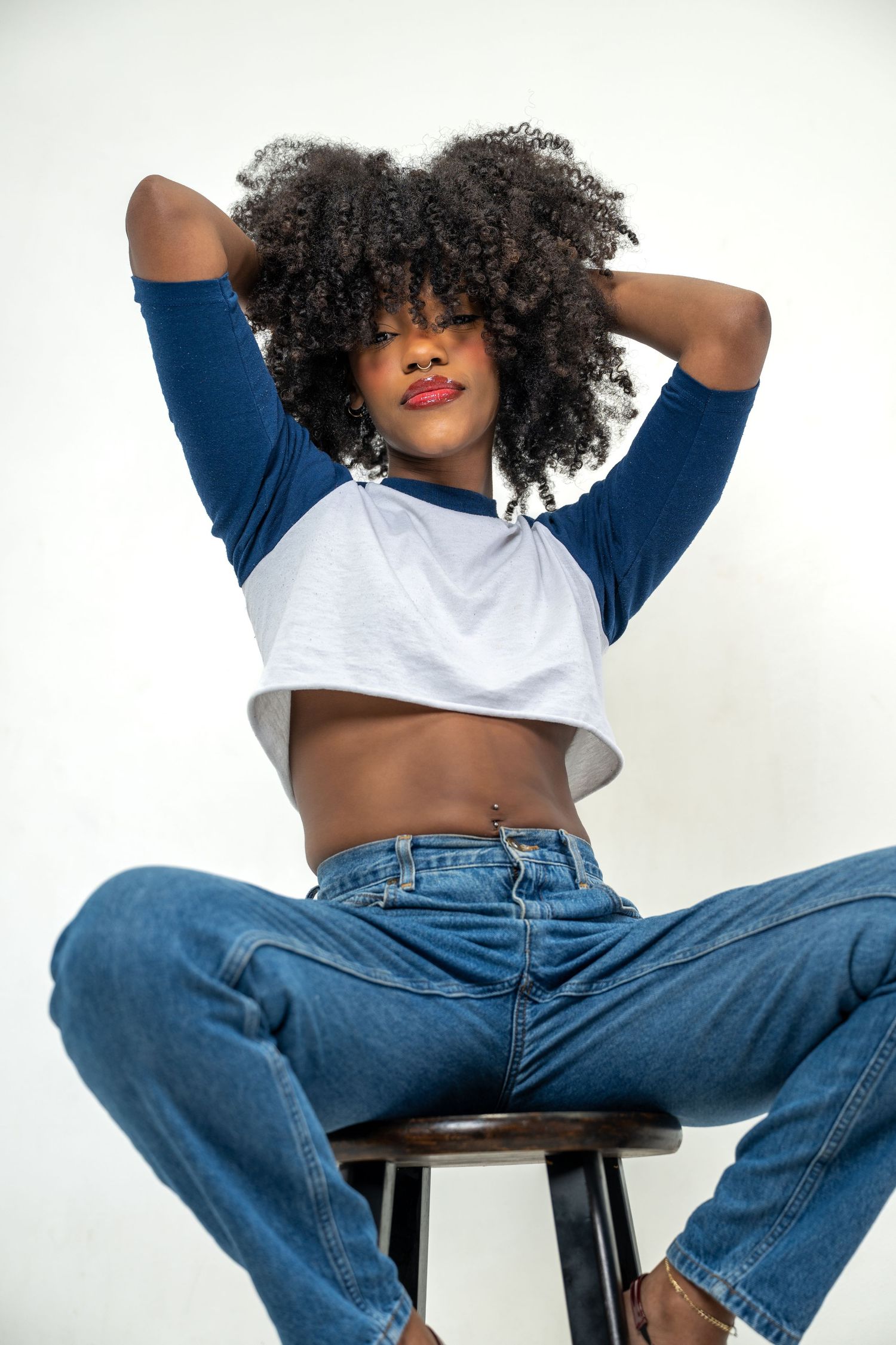 Young Black woman with curly afro sits on stool, wearing crop top and jeans, arms raised behind head, white background.