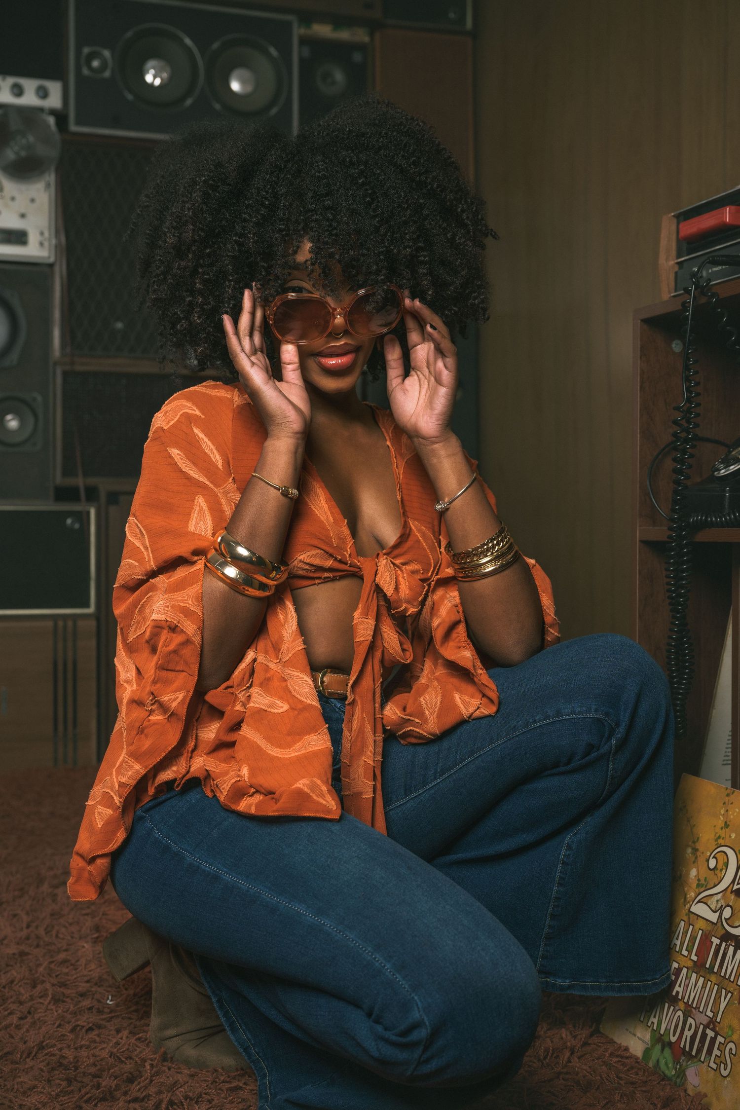 A woman with an afro in an orange blouse and wide-leg jeans adjusts oversized sunglasses in a retro music room.