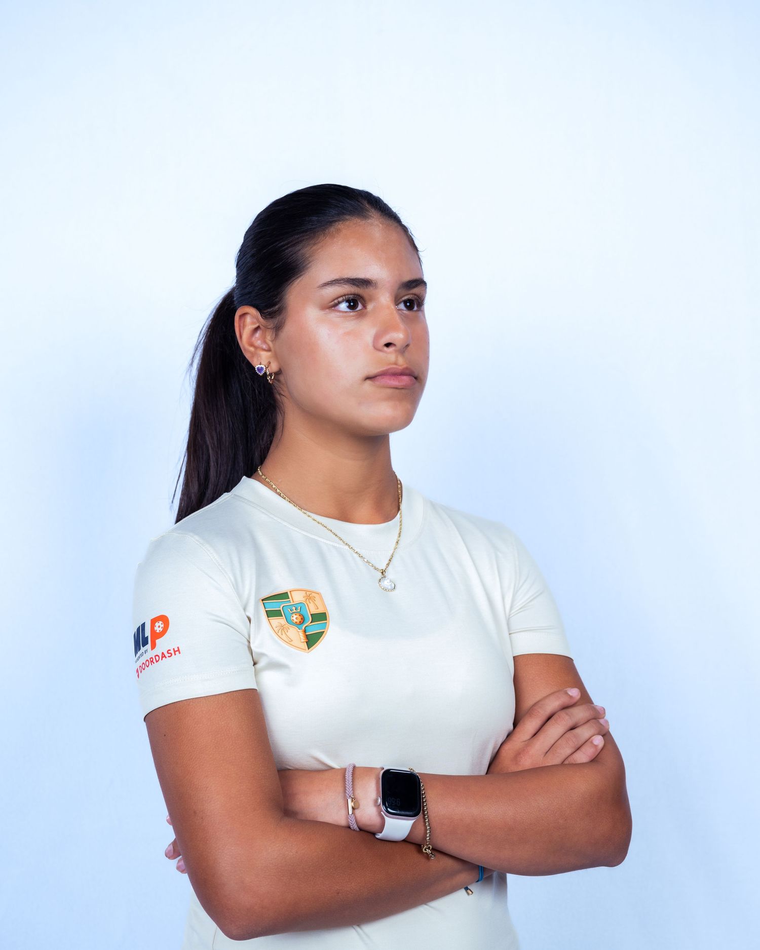 Young woman in white sports shirt with arms crossed, wearing a smartwatch, posing against a light blue background.