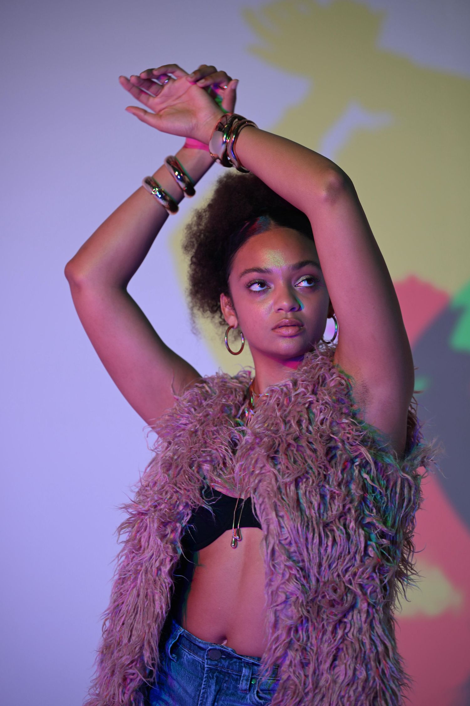 Black woman in fur vest raises arms gracefully under colorful neon lighting with shadow projection.