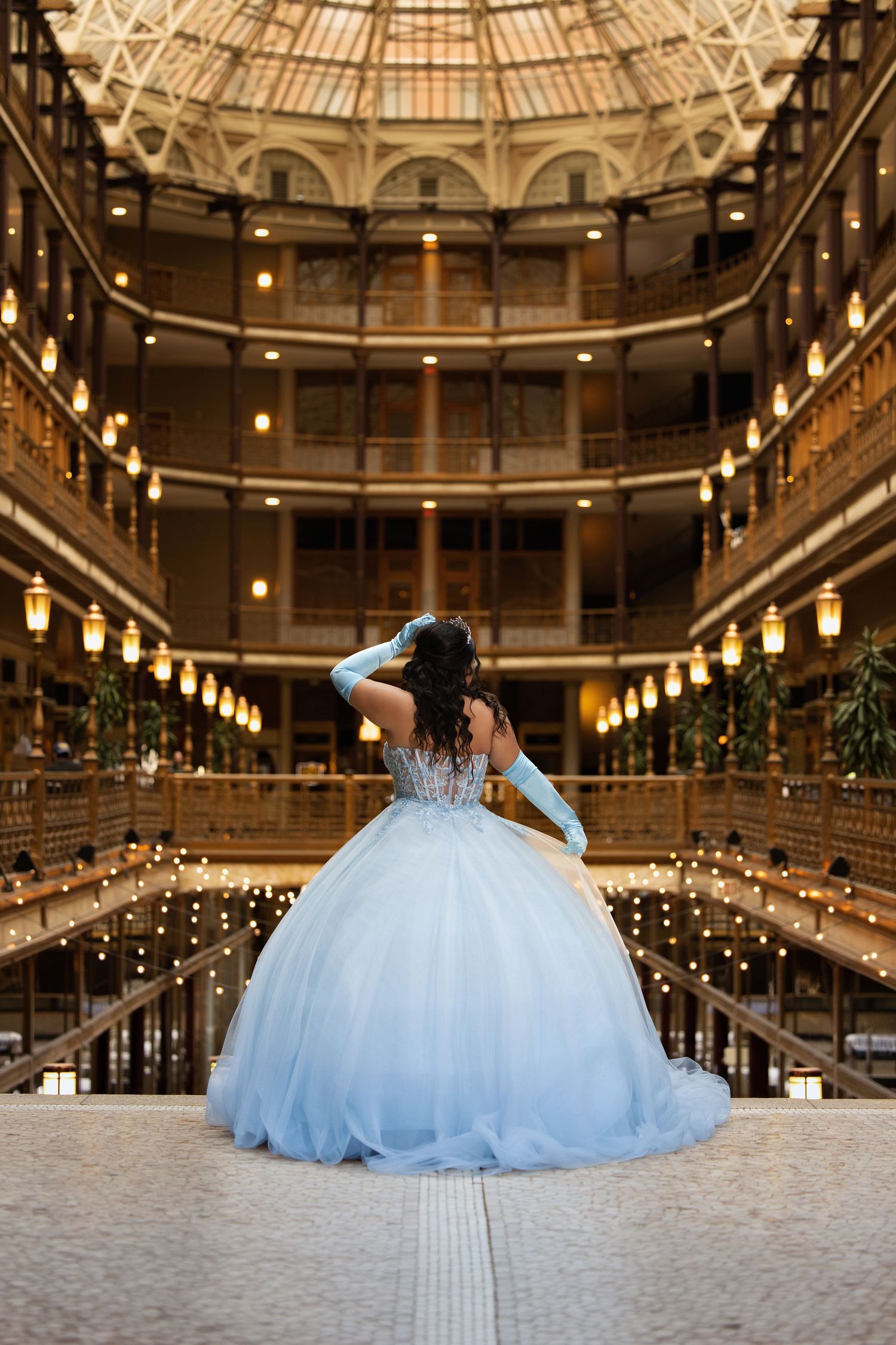 Woman in light blue ball gown with gloves poses in ornate multi-story atrium with warm glowing lights.