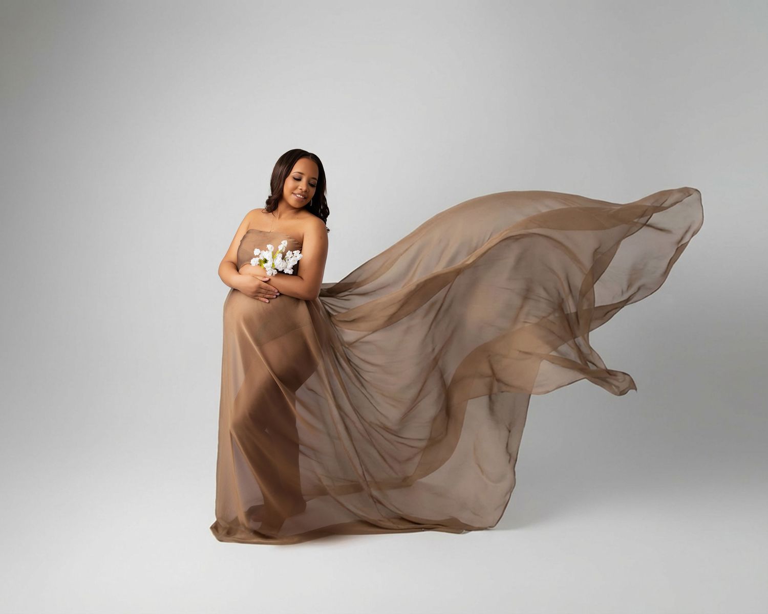Pregnant woman in flowing brown chiffon gown holding flowers, elegant maternity portrait on white background.