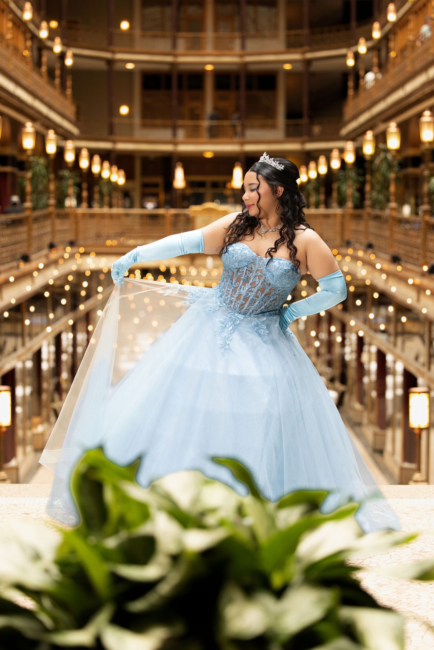 Young woman in a light blue quinceañera ball gown with tiara and gloves poses in an elegant golden-lit venue.