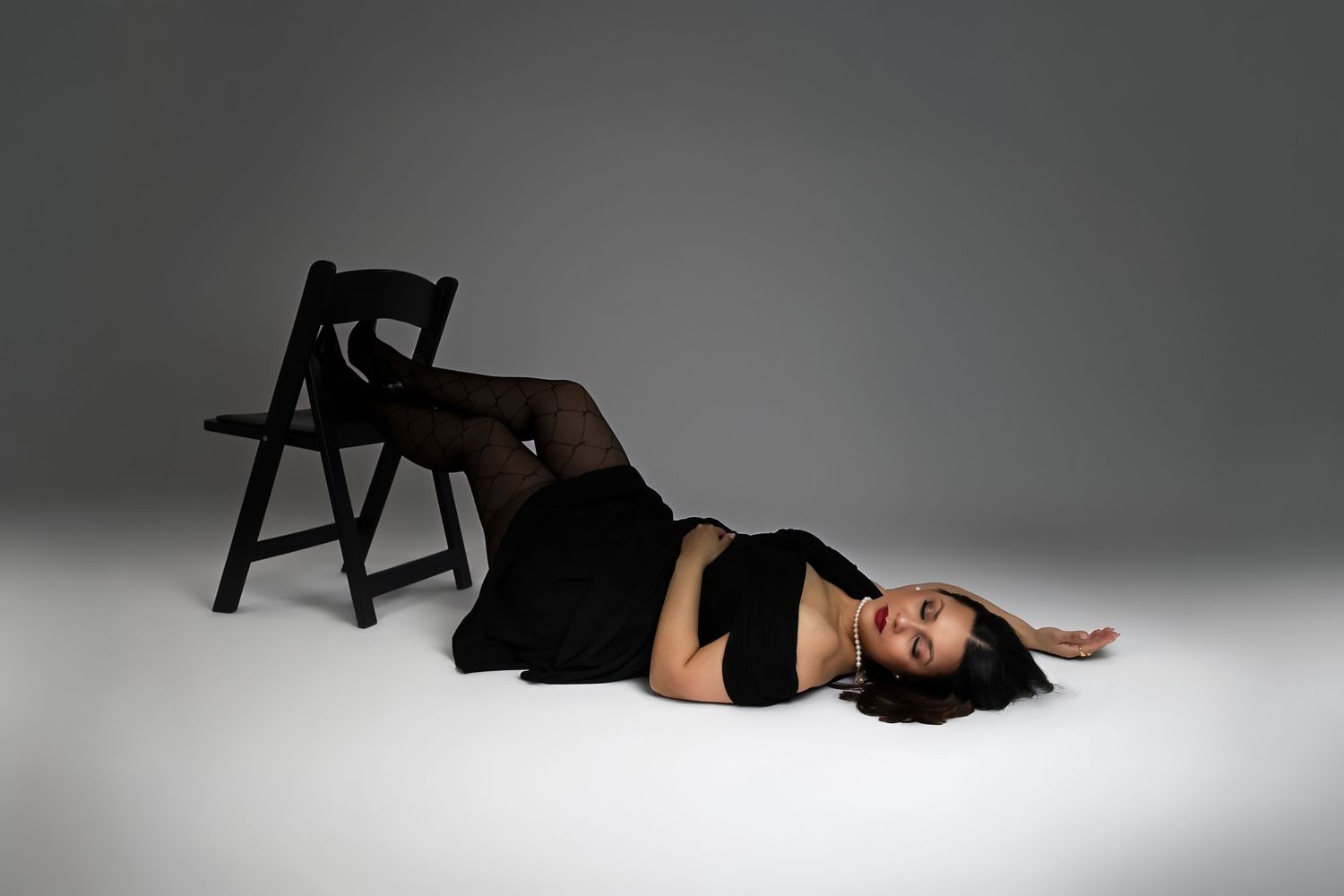 Woman in black dress and tights lying on studio floor beside a black folding chair against gray background.