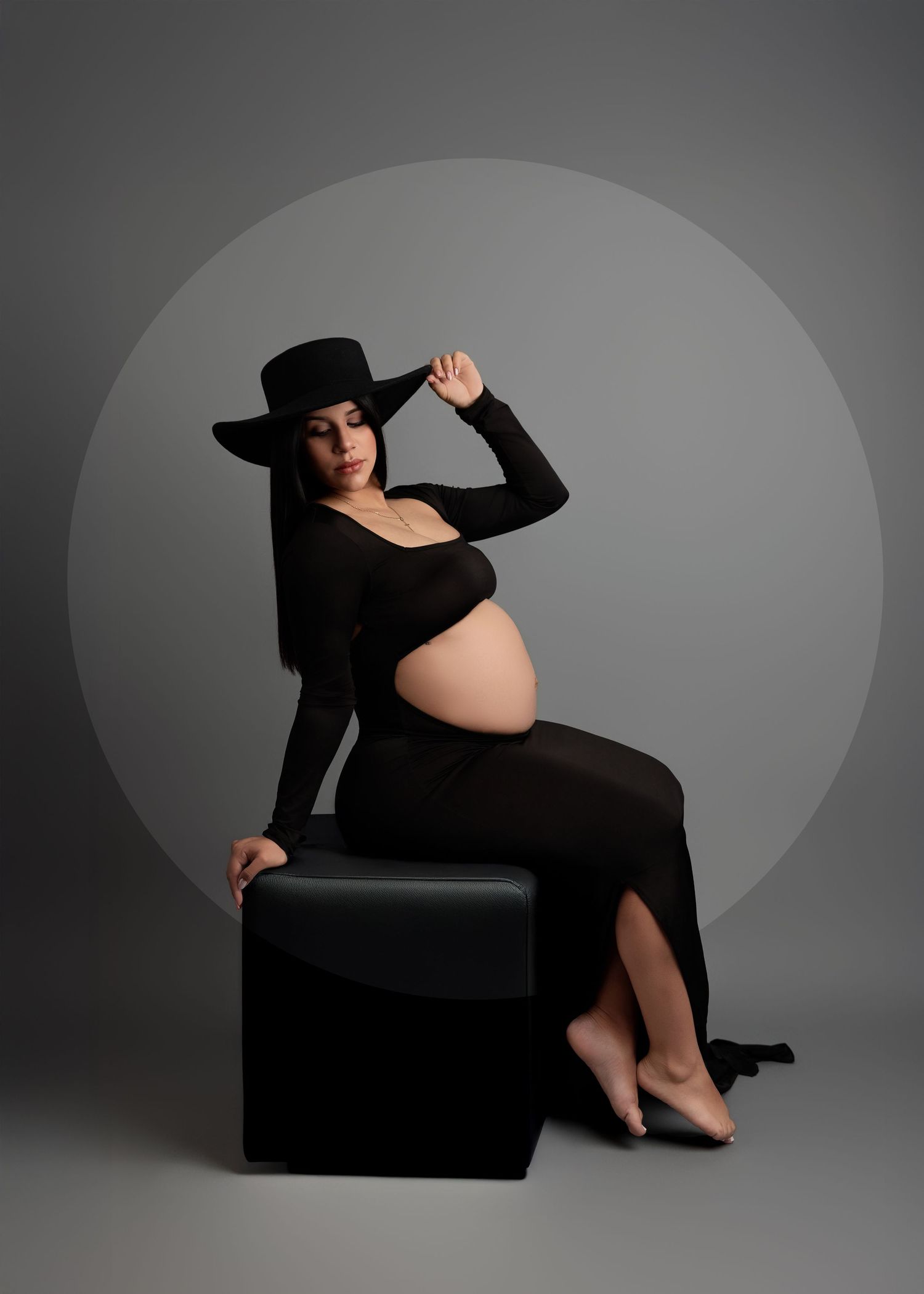 Artistic maternity photoshoot with figure in black dress and hat posing dramatically against dark gray background.