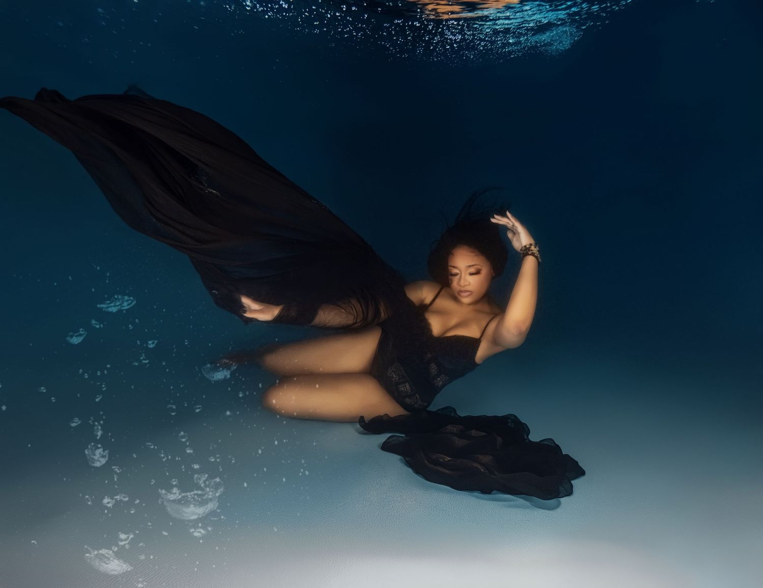 Underwater dancer in black dress moves gracefully through deep blue water with bubbles trailing behind.