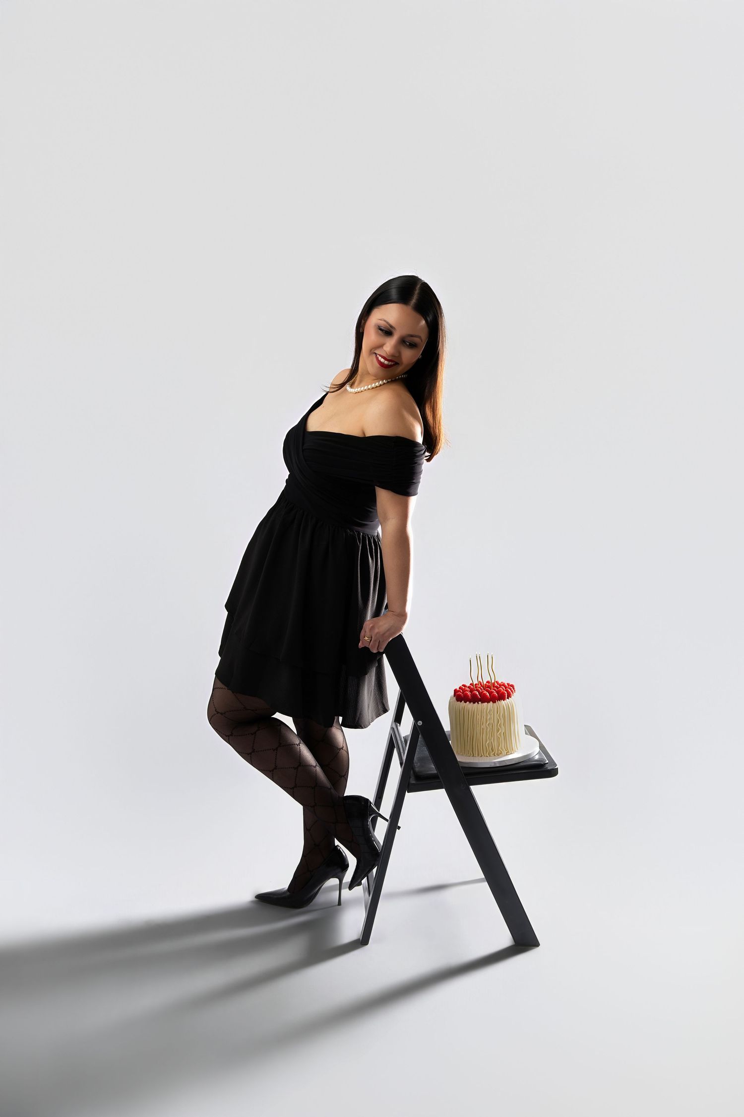 Woman in black off-shoulder dress stands beside a ladder shelf holding a birthday cake with candles.