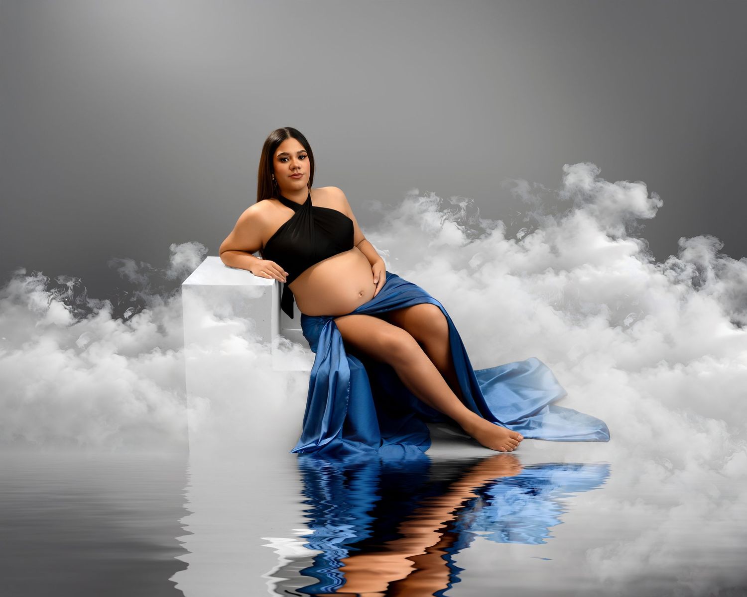 Maternity photo with flowing blue fabric against dramatic clouds and water reflection in black and white background.