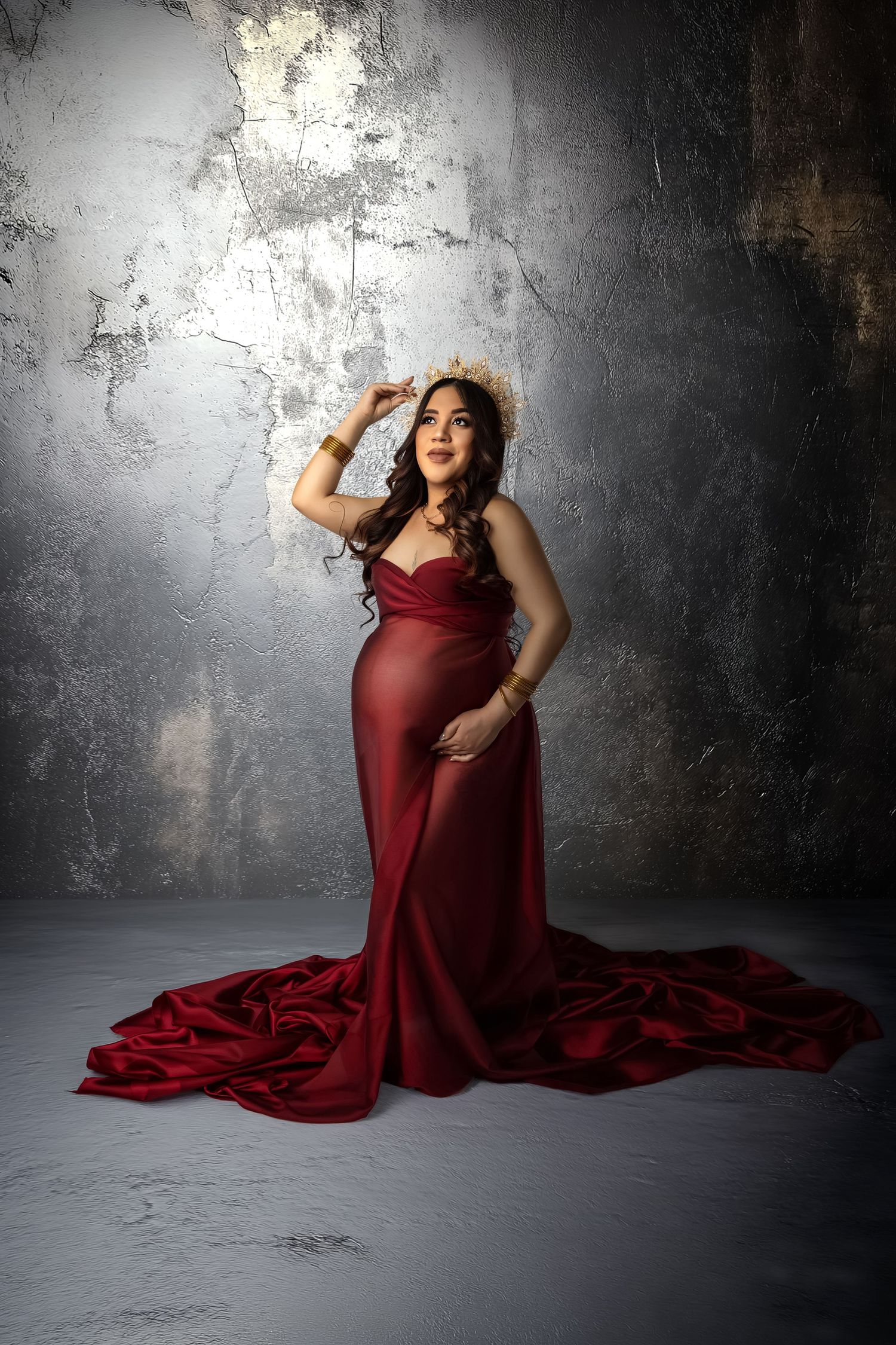 A pregnant woman poses in a flowing burgundy gown against a textured gray wall in dramatic studio lighting.