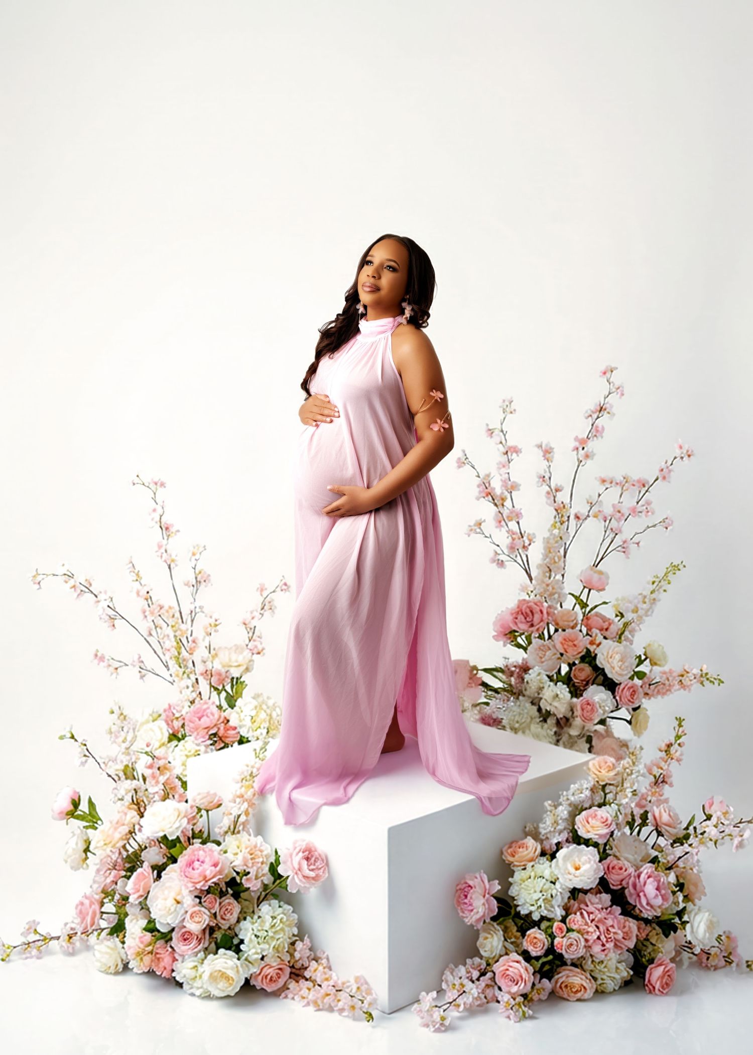 Pregnant woman in pink halter gown stands on white cube surrounded by pink and white floral arrangements in studio.