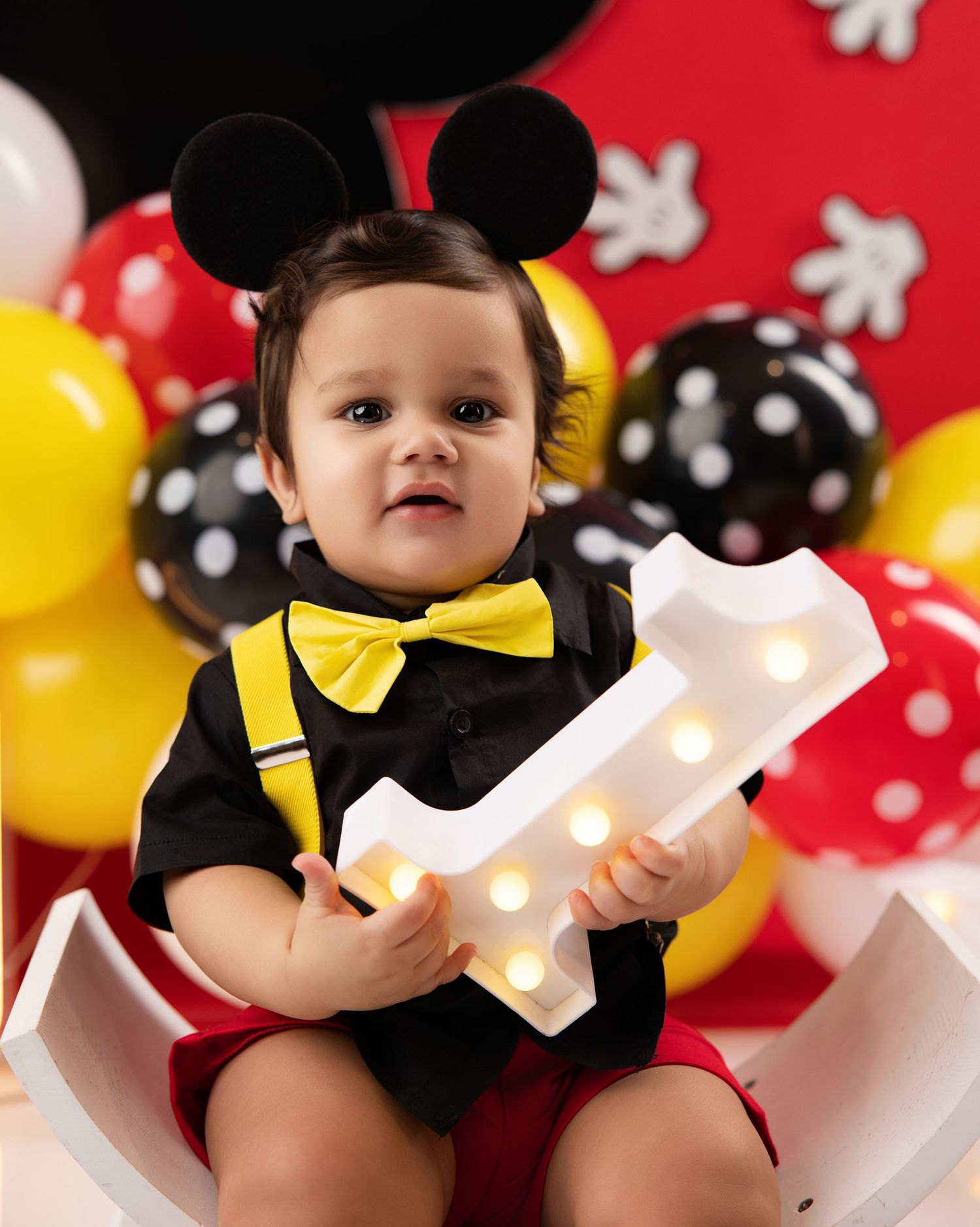 Baby in Mickey Mouse ears holds a lit number 1 sign at a first birthday party with colorful balloons.