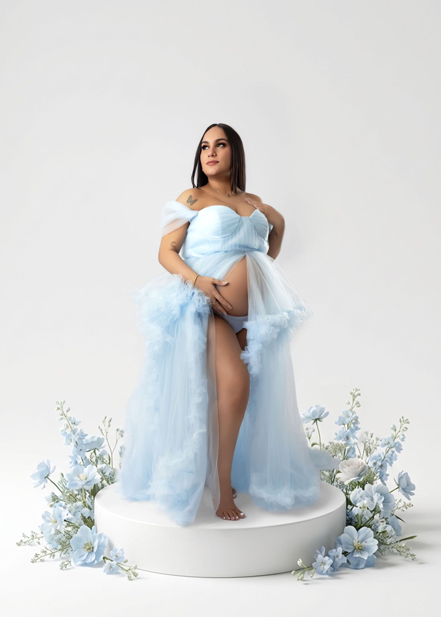 Maternity photo session featuring light blue tulle gown and delicate floral decorations on white background.